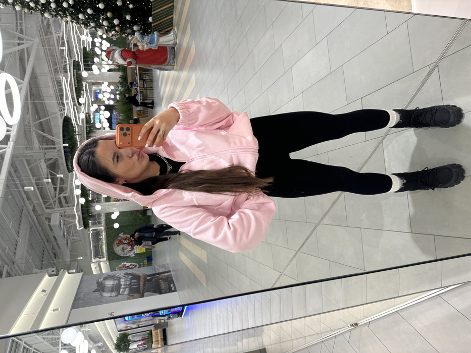 A forensic description of the image reveals a young adult female posing for a mirror selfie within an indoor commercial establishment, specifically a shopping mall or large retail space in Yudino, Russia.
**Subject:** The primary subject is a female, appearing in her 20s to 30s, with long dark brown hair parted down the middle and wearing a light pink hooded jacket, black form-fitting leggings, and black chunky boots with visible white socks. A silver chain necklace with a triangular pendant is visible around her neck. She holds an orange-cased smartphone in her right hand, positioned to capture her reflection, while her left hand is placed in the jacket pocket. Her facial expression shows pursed lips directed towards the phone.
**Environment and Background:** The setting is a brightly lit, spacious indoor area with a light gray tiled floor. The high ceiling features exposed white structural elements and numerous modern lighting fixtures, including circular and linear LED lights. To the subject's right (viewer's left in the reflection), a large, decorated Christmas tree with white ornaments and lights stands next to a life-sized Santa Claus figure and another smaller decorative figure, likely a Snow Maiden. Beyond these decorations, tables and chairs indicate a dining or seating area. To the subject's left (viewer's right in the reflection), a large graphic advertisement featuring plaid blankets is visible on a wall. Several indistinct figures are walking in the distant background, appearing out of focus, one wearing a dark jacket and walking away. The overall environment suggests a bustling public space adorned with holiday decor.