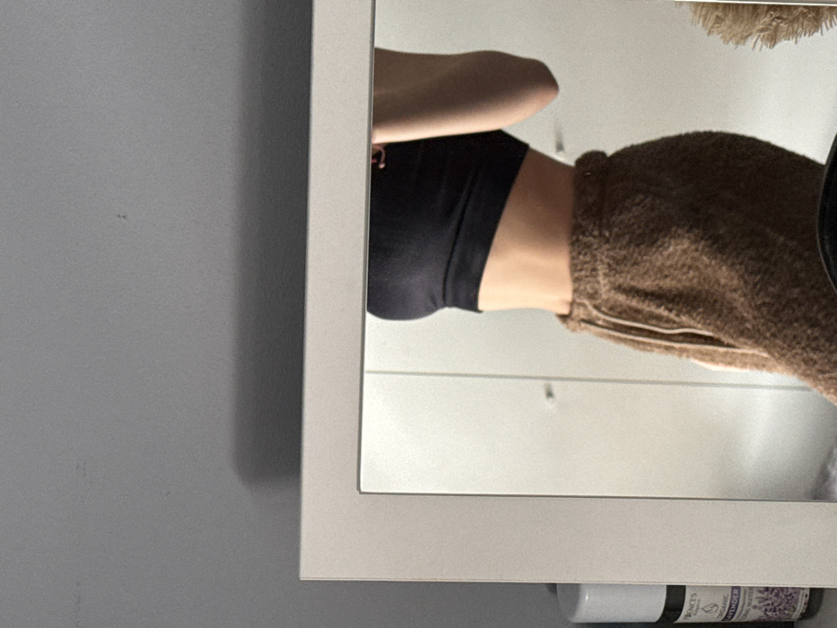 The image depicts a partial reflection of a female individual's torso in a rectangular mirror with a white frame. The person is wearing a black crop top or sports bra and textured, brown sweatpants with a light-colored drawstring. The reflection shows the individual from the mid-chest to the upper thighs, observed from a slight left-side profile.
Reflected in the mirror's background are white, modular wardrobe or closet doors. To the far left, a white cylindrical container and a dark glass bottle are visible. The bottle's label clearly displays "PRONCES" and "ORGANIC LAVENDER" in English, along with "БЪЛГАРСКО МАСЛО ОТ ЛАВАНДУЛА" in Cyrillic, identifying it as Bulgarian Lavender Oil. This product indicates the scene is set within a residential interior in Bulgaria.
The environment outside the mirror consists of a plain, light gray wall in the upper portion of the image. A partial view of a white cylindrical object, likely the same one reflected, is seen on the left side, adjacent to the mirror frame. The individual appears to be taking a self-portrait, holding a device to capture their reflection. Based on the presence of the Bulgarian product and the explicit instruction for location, the scene is situated within a residential interior in Sofia, Bulgaria.