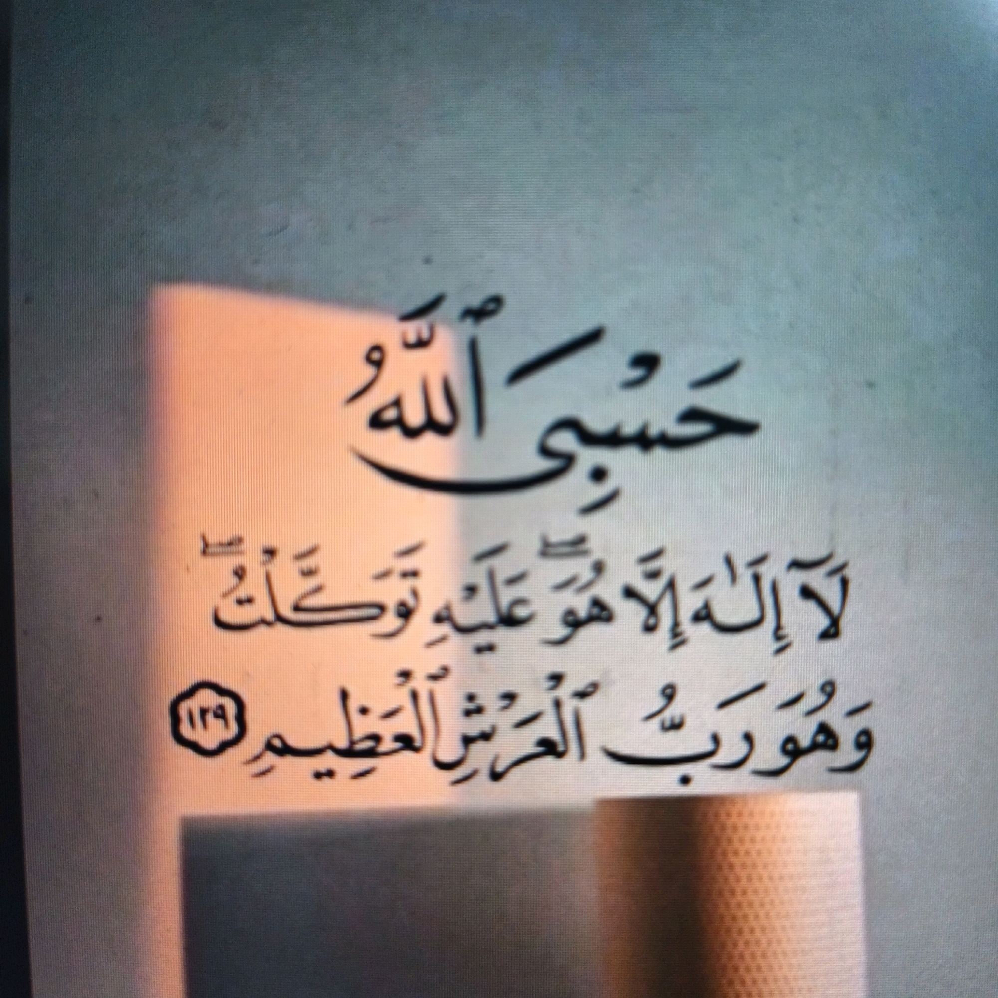 The image is a close-up shot of Arabic calligraphy written on a page. The calligraphy appears to be from a religious text, likely the Quran, given the subject matter. The first line reads "حَسْبِيَ اللَّهُ" (Hasbi Allah), which translates to "Allah is sufficient for me." The second line reads "لَا إِلَٰهَ إِلَّا هُوَ ۚ عَلَيْهِ تَوَكَّلْتُ" (La ilaha illa huwa 'alayhi tawakkaltu), meaning "There is no god but He; on Him I rely." The third line reads "وَهُوَ رَبُّ الْعَرْشِ الْعَظِيمِ" (Wa huwa Rabb al-'arsh al-'adhim), which translates to "And He is the Lord of the Great Throne." A circular emblem containing the number "129" is visible to the left of the third line. The text is written in black ink on a light-colored background. A warm light source illuminates the left side of the image, casting shadows and creating a gradient effect on the page. The background of the image is a muted, textured surface, possibly a wall or a fabric. The image quality is somewhat blurry, suggesting it was taken with a phone camera at a distance or in low light conditions. There are no people or specific landmarks visible in the image. The city of Poonch, India, is not depicted or suggested in this image.