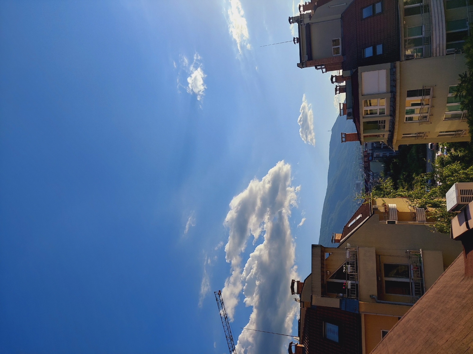 The image is a vertical, wide-angle shot of buildings and a bright blue sky with scattered clouds, taken from a high vantage point. The perspective is tilted, suggesting the photo was taken looking upwards or from a sloped surface. In the foreground, the roofs of several buildings are visible, featuring terracotta tiles and modern architectural elements like balconies and window frames.
Further back, apartment buildings with light-colored facades and darker tiled roofs line a hillside. Some buildings have visible air conditioning units mounted on their exteriors and balconies with railings. The architecture suggests an urban environment.
Dominating the upper left of the frame is a vast expanse of clear blue sky, streaked with thin, wispy clouds and illuminated by bright sunlight, indicating it is daytime. In the distance, a mountain range is visible, partially obscured by trees and buildings. A construction crane, with its red and white lattice structure, stands prominently against the sky, suggesting ongoing development or renovation.
The overall mood is bright and summery, with the clear sky and sunlight creating a cheerful atmosphere. The presence of the crane indicates an urban setting undergoing development, common in a city like Sofia, Bulgaria. No visible text is present in the image.