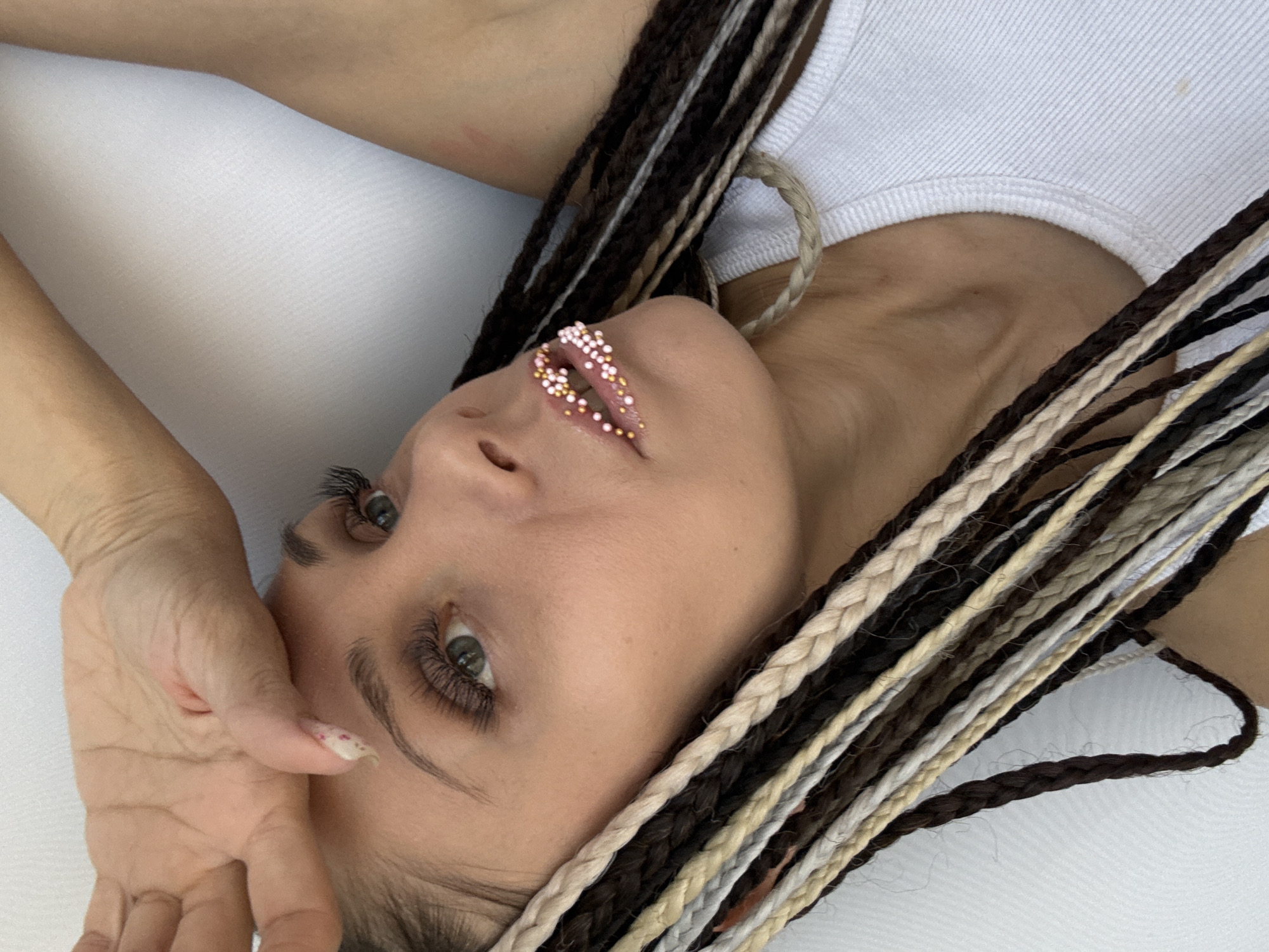 A woman with braided hair and beaded lips is lying on a white surface. She is wearing a white tank top. Her right hand is raised to her face. The location is Unknown, Unknown.