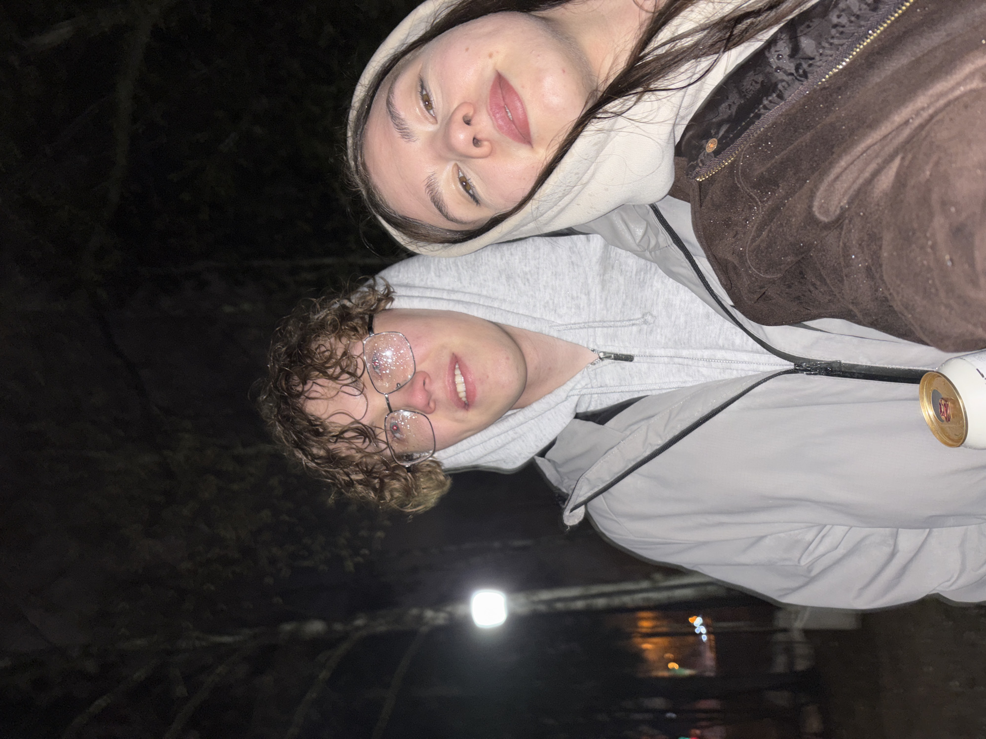 Two people are in the image at night. One is a fair-skinned person with dark hair and is wearing a white hoodie and a brown jacket. The other person, who appears to be fair-skinned, is wearing bedazzled glasses and a light grey hoodie and jacket and has curly hair. They are standing outside in front of trees with some buildings blurred in the background. The photo was taken at night in Perm, Russia. A street light is also in the picture.