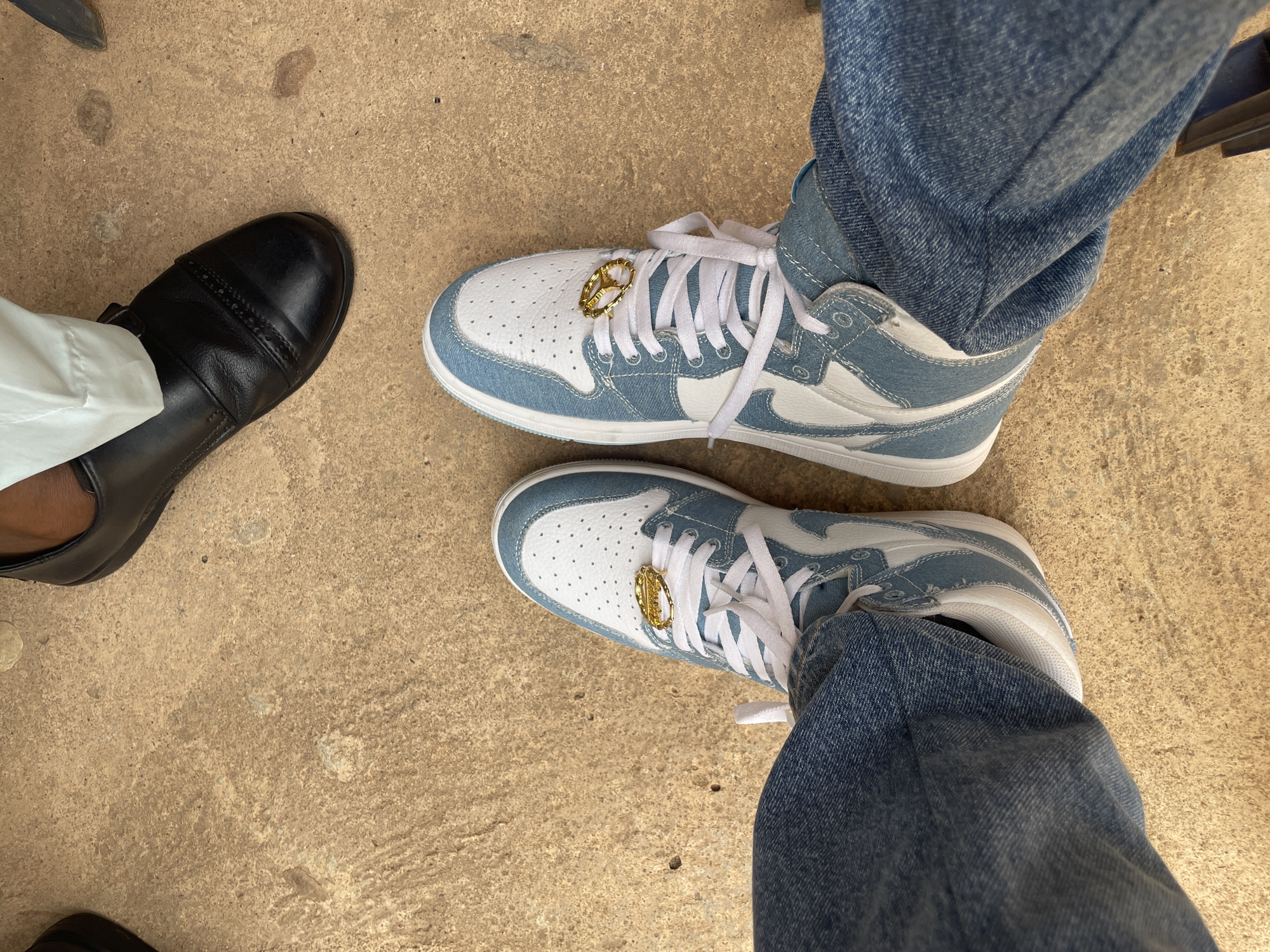 A low-angle, eye-level shot shows two pairs of shoes on a concrete surface. On the left is a black leather dress shoe with a white cuff above. On the right are two blue and white sneakers with gold lace charms. One pair of the shoes is being worn with blue jeans. The location appears to be in Akwanga, Nigeria.