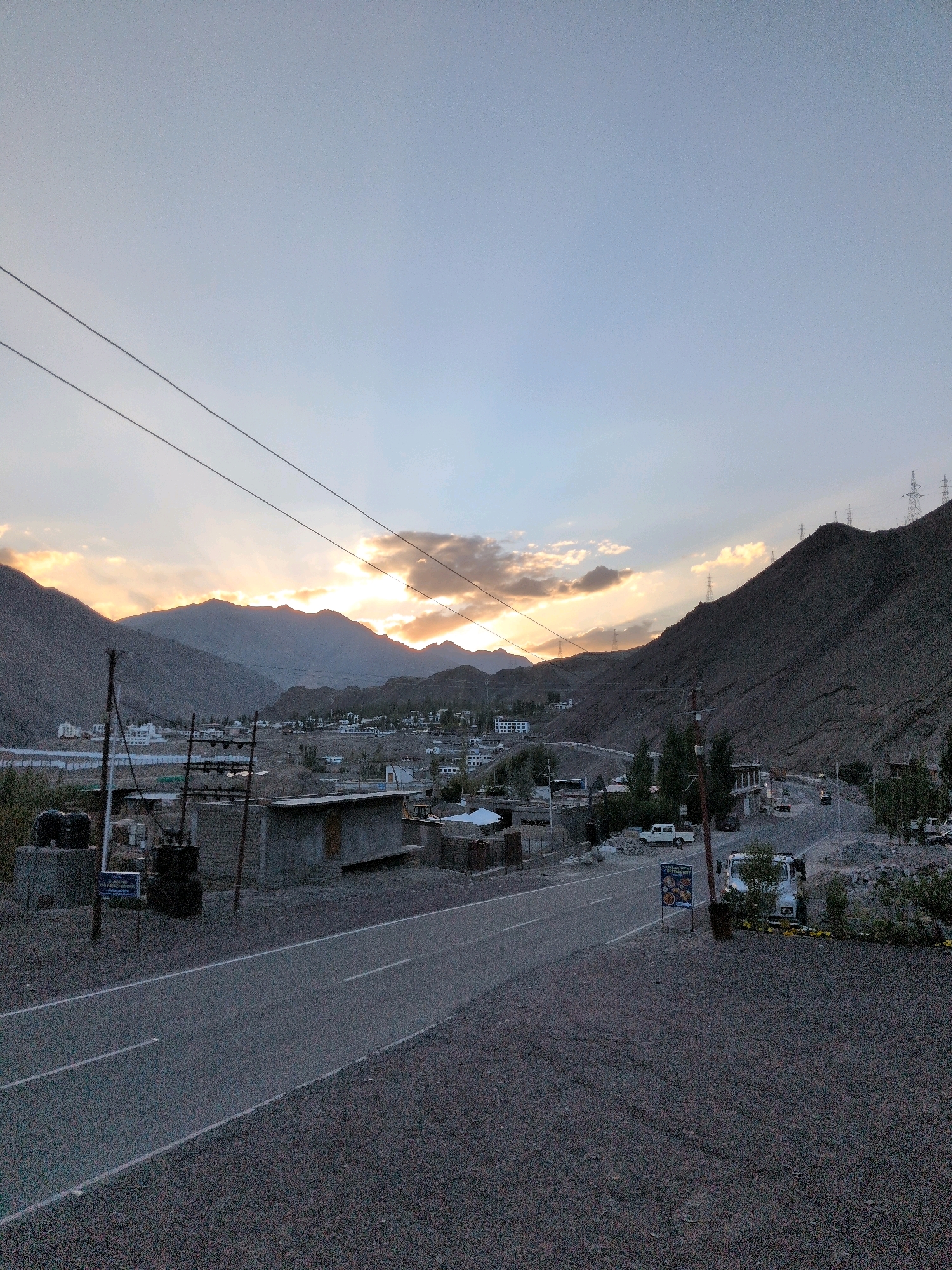 The image depicts a rural landscape in Khaltse, India, during what appears to be twilight or dawn. A paved road, marked with white lines, cuts through the foreground, leading towards a valley filled with scattered buildings and structures. The background is dominated by imposing, barren mountains, their slopes etched with shadows. The sky above is a gradient of soft blue and muted orange, with wispy clouds catching the last or first light of the sun. Several power lines stretch across the frame, supported by poles.
In the mid-ground, a small settlement is visible, characterized by low-rise buildings and some construction activity. A white pickup truck is parked on the side of the road, and further down, another vehicle is visible. There are a few trees scattered throughout the scene, adding touches of green to the predominantly earthy tones. A signpost, likely for a restaurant, stands near the roadside in the right foreground. The overall impression is one of a quiet, remote location bathed in soft, diffused light. There are no visible people in the image.