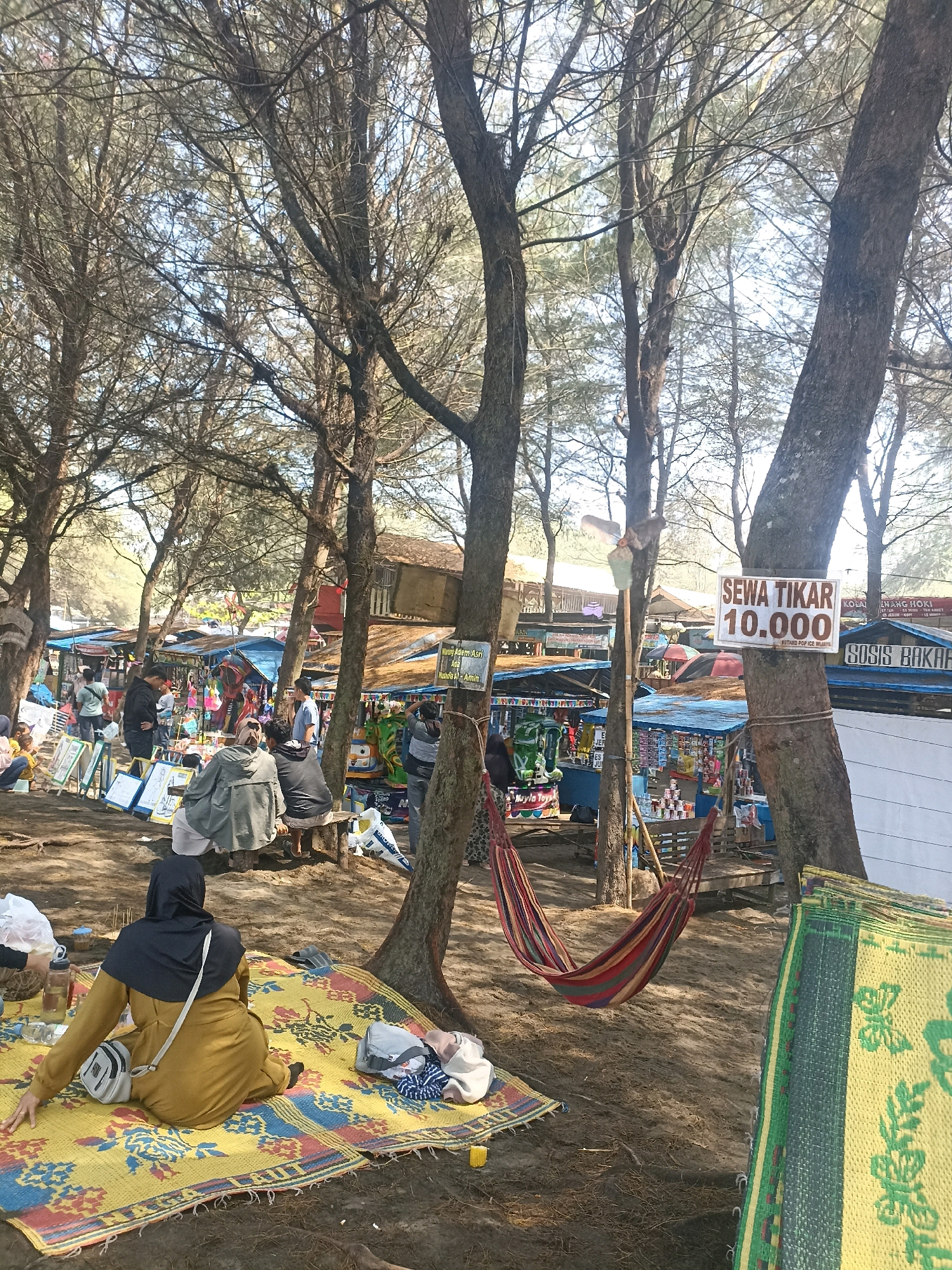 The image depicts a busy outdoor market area at a beach in Kenoyajaya, Indonesia. Several large trees with bare branches provide shade over the sandy ground where numerous stalls are set up. The stalls appear to be selling various goods, including food, drinks, and possibly souvenirs, indicated by the colorful displays and awnings. A prominent sign on a tree trunk reads "SEWA TIKAR 10.000," suggesting that mats are available for rent at 10,000 Indonesian Rupiah. People are seen browsing the stalls, sitting on benches, and relaxing on mats. In the foreground, a person wearing a yellow hijab and dress is sitting on a colorful woven mat. Further back, a hammock is strung between two trees, adding to the relaxed atmosphere. The overall scene suggests a lively and active marketplace, likely catering to beachgoers.