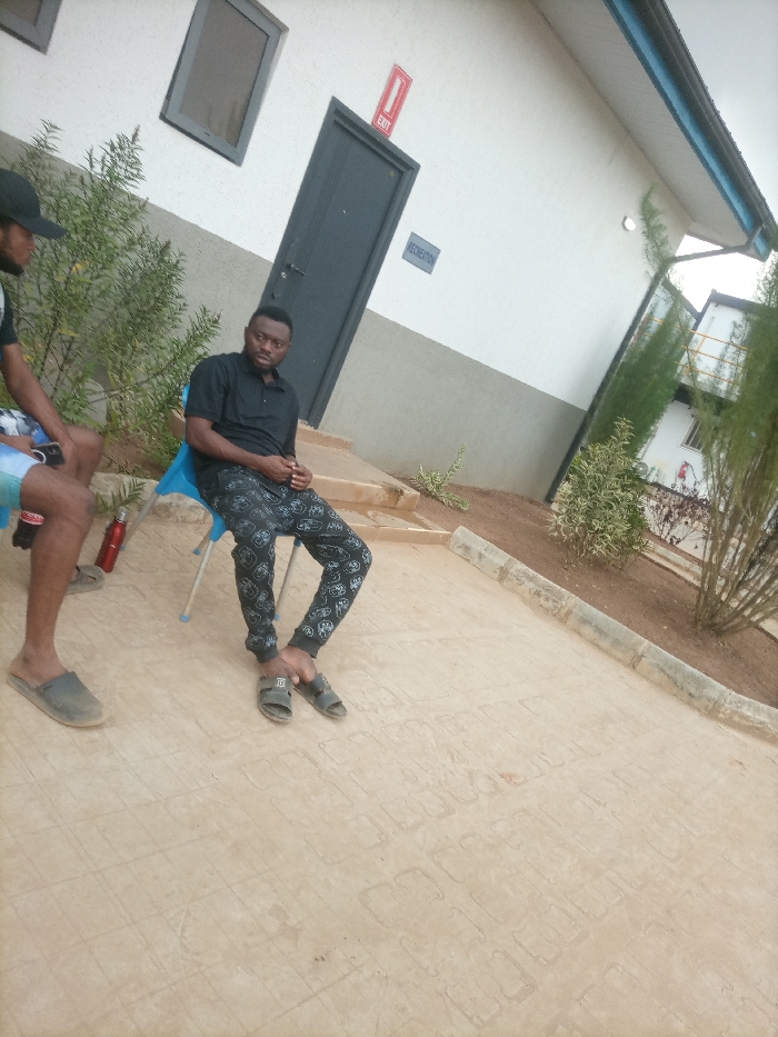 Two men are seated outside a building with a grey door labeled "Exit" and "Recreation." One man wears a black shirt and patterned pants, sitting on a blue chair. The other man, partially visible, wears a black cap and patterned shorts. A red water bottle sits on the ground nearby. The building is white with a grey base and window. Green bushes and trees line the area. The ground is paved with a textured pattern.