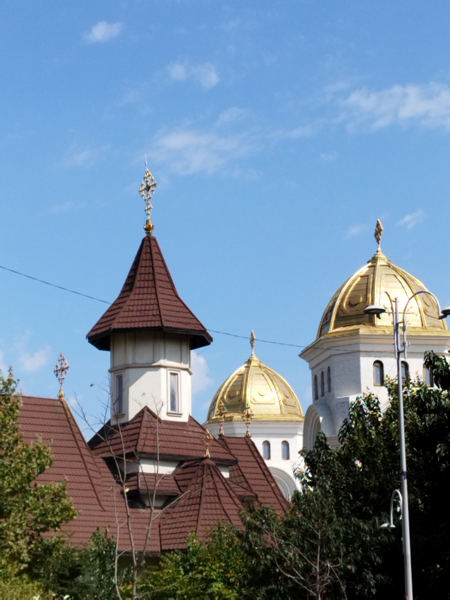 The image shows the exterior of the Saint George's Church in Bucharest, Romania, on a clear day with a blue sky and scattered white clouds. The church features multiple domes, some of which are golden and topped with crosses, and others are conical with brown tiled roofs. A prominent white tower with a conical roof and a cross is visible on the left side of the frame. Lush green trees surround the lower parts of the building. The scene is static and depicts the architecture of the church against a natural background.