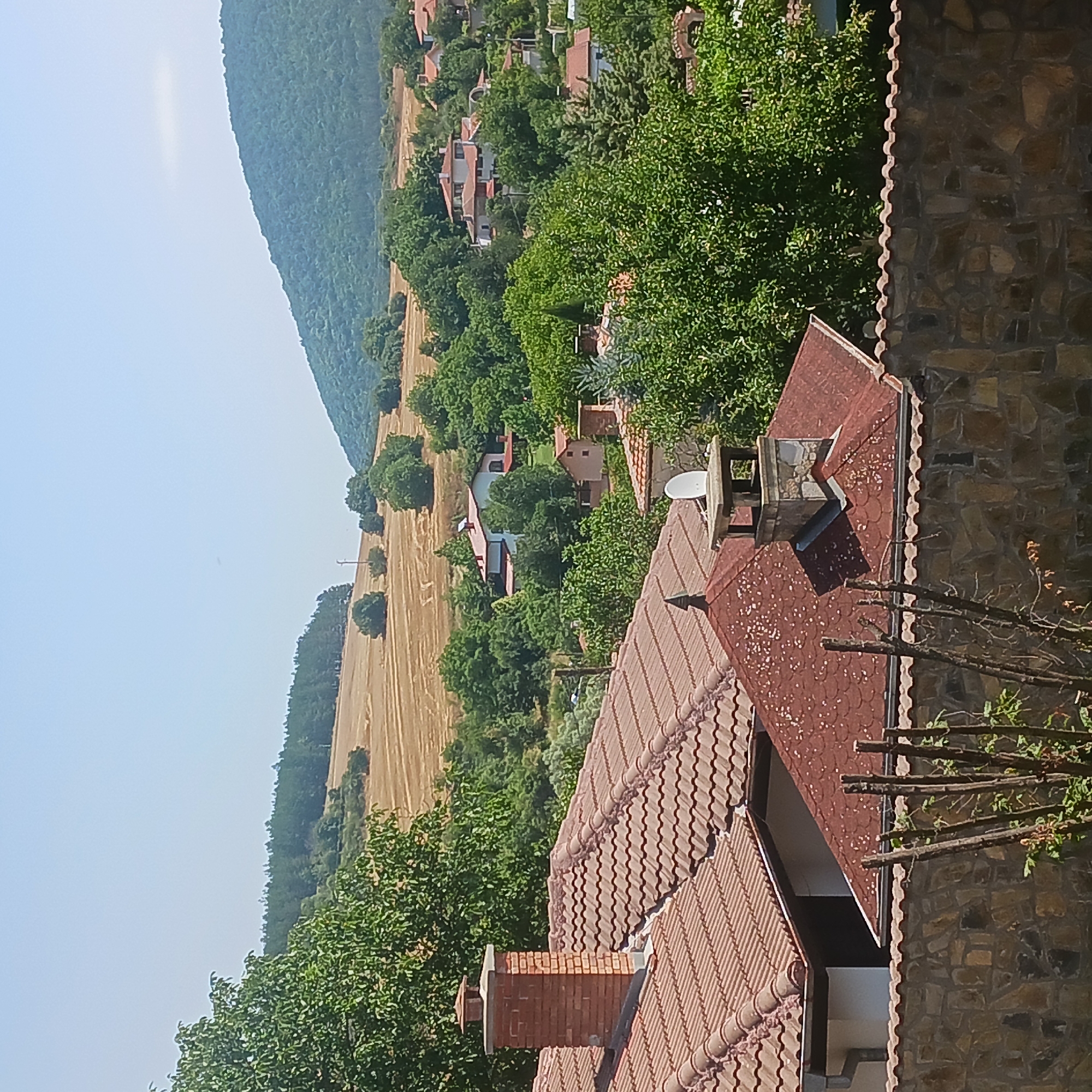 The image depicts a panoramic view of the village of Sulitsa, Bulgaria, on a clear, sunny day. The foreground features a stone wall with weathered wooden posts, suggesting a boundary or fence. Above the wall, several tiled roofs of houses are visible, two of which have prominent brick chimneys. One chimney on the left is made of red brick and has a decorative cap, while another on the right is constructed with stone and features a satellite dish mounted on the adjacent rooftop.
The middle ground is characterized by lush green trees and more houses nestled amongst them, their roofs also predominantly tiled. A white house with a red-tiled roof stands out amidst the foliage. In the background, a vast, golden field, likely a harvested crop, stretches towards a densely wooded hill and a larger, verdant mountain that dominates the skyline. The sky is a pale blue, with a slight haze. The overall scene conveys a sense of rural tranquility.