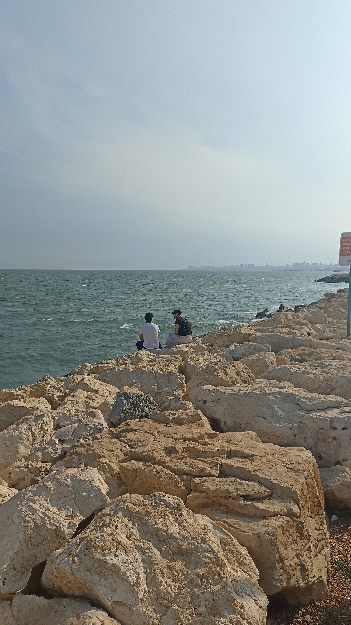 Two individuals are seated on a rocky shoreline, facing a body of water. The person on the left is wearing a white t-shirt and dark pants, while the person on the right is wearing a black t-shirt and a dark cap. They are situated on a breakwater composed of large, light-colored rocks. The sea is choppy with visible waves, and a hazy skyline of a city is visible in the distance. The sky is mostly clear with some faint clouds. A sign is partially visible on a pole to the right of the image, indicating a warning or information. This scene appears to be located on the coast of Yenişehir, Turkey, a region known for its coastal areas and urban developments along the water.