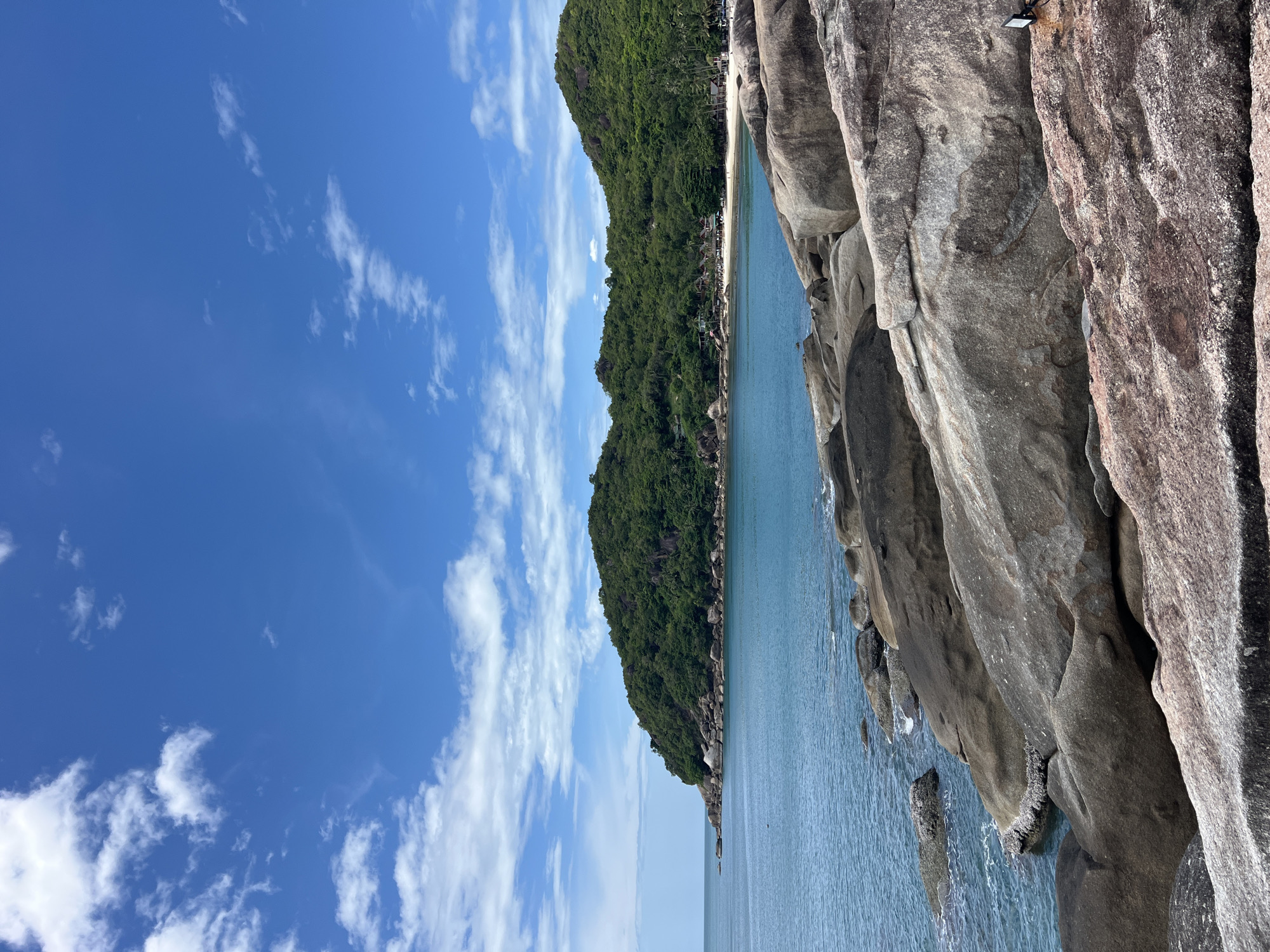 This image depicts a tropical coastal landscape in Baan Chaweng Noi, Thailand, under a bright blue sky with scattered white cumulus clouds. The scene is dominated by a calm, clear turquoise-blue sea that occupies the mid-ground.
On the left, a rocky headland covered densely with lush green tropical vegetation extends into the water. To the right, another larger, heavily wooded landmass slopes down to reveal a crescent-shaped, light-colored sandy beach. Along this beach, several low-rise structures, likely resort buildings or bungalows, are visible amidst the trees, and multiple small, indistinct human figures are present on the sand.
The foreground is characterized by large, smooth, weathered boulders and rock formations, ranging in color from dark grey to reddish-brown. Some of these rocks on the left are partially submerged in the water, exhibiting white marine growth along the waterline. In the lower right corner, resting on a reddish-brown rock, a small black rectangular object, possibly a light fixture or solar panel, is visible.