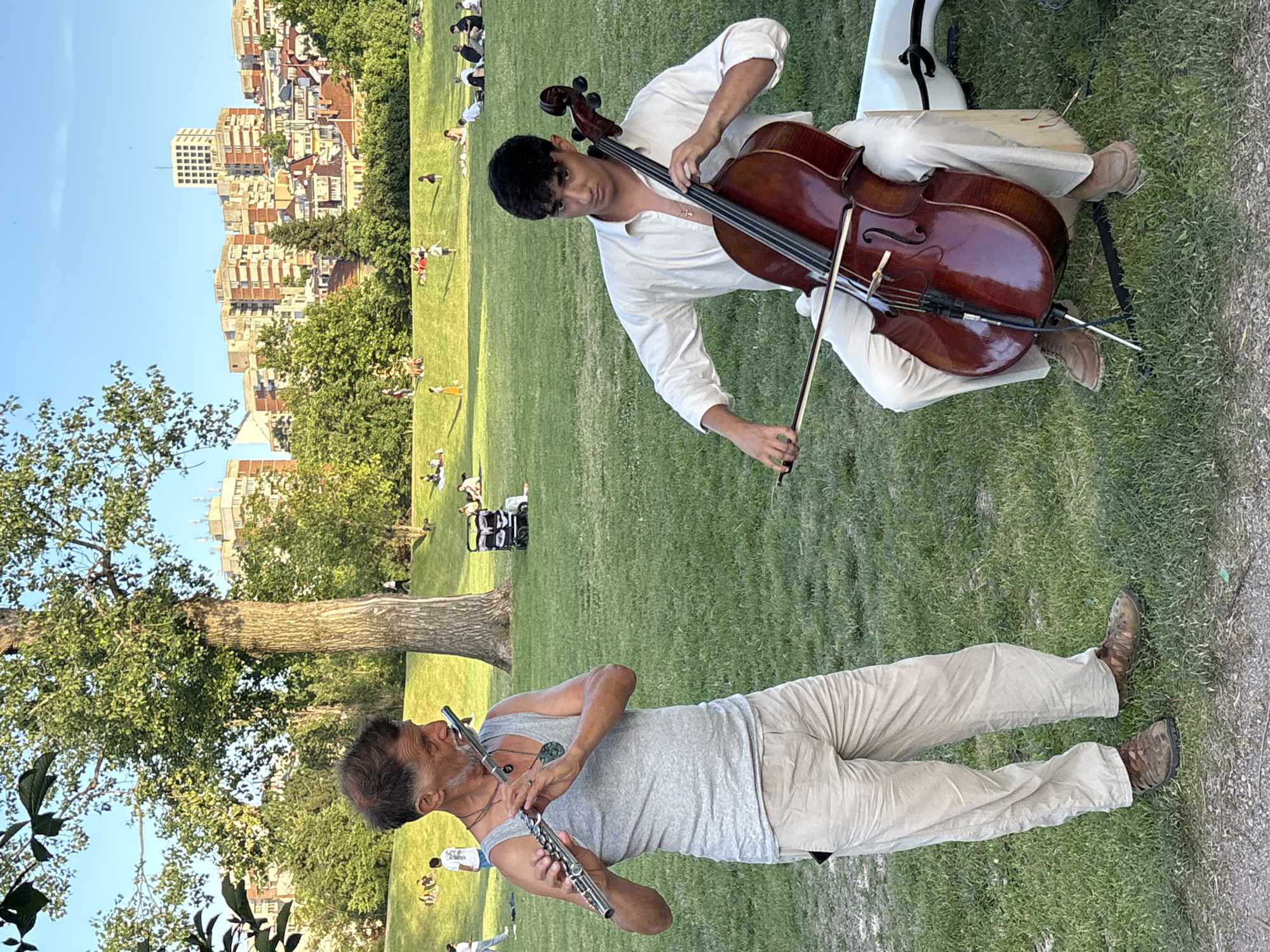 Two men are playing musical instruments in a park in Sofia, Bulgaria. On the left, an older man with gray hair, wearing a gray tank top and beige pants, plays a silver flute. He is standing and looking to his left. On the right, a younger man with dark hair, wearing a white button-down shirt and beige pants, sits and plays a cello. The cello is a deep red-brown color. The men are facing away from each other, and the cello player is looking towards the camera. The park has a lush green lawn, and in the background, there are trees and buildings characteristic of Sofia. Several other people are visible in the background, some sitting on the grass and others walking. The sky is clear and blue, suggesting it is a sunny day.