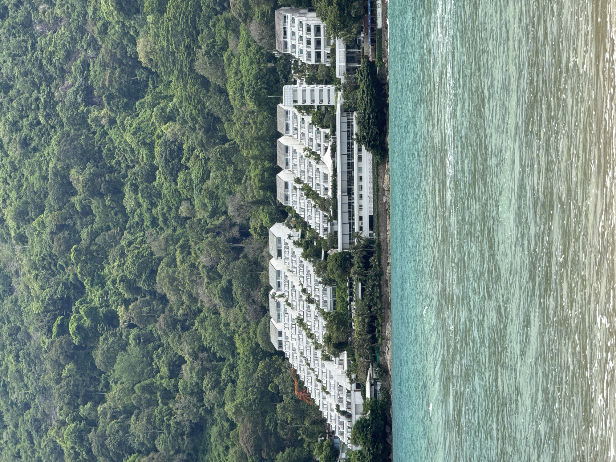 A wide shot captures a resort built into a lush, green hillside overlooking a turquoise ocean. The resort buildings are white and tiered, resembling a staircase ascending the slope. The dense jungle foliage covers the upper portion of the image. The water in the foreground is calm with gentle waves and some white foam near the shore. The scene is bathed in natural daylight, suggesting it is daytime. There are no people visible in the image.