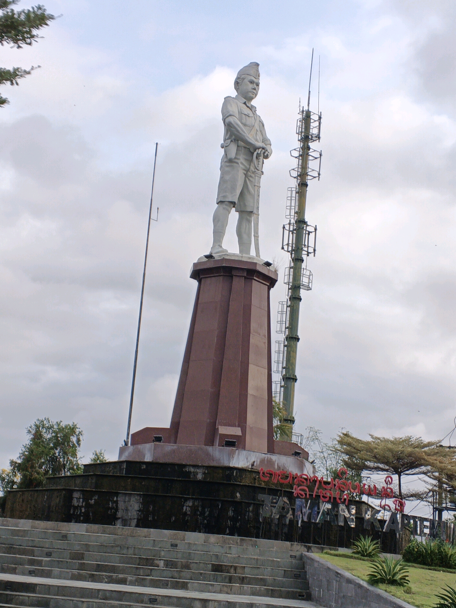 The image shows a monument with a statue of a man in military attire standing on a tall, segmented, dark red pedestal. To the right of the statue, a tall, multi-tiered communication tower is visible. In front of the pedestal, there is a tiered structure with dark, weathered stone work. A large set of concrete stairs leads up to this tiered structure. Behind the monument and tiered structure, there is signage that appears to be in a foreign language, with large red letters and metallic block letters spelling out "TAMANKARIM". Lush greenery, including trees and bushes, is present around the monument and stairs. The sky is cloudy. The monument is located in the city of Blahbatuh, Indonesia.