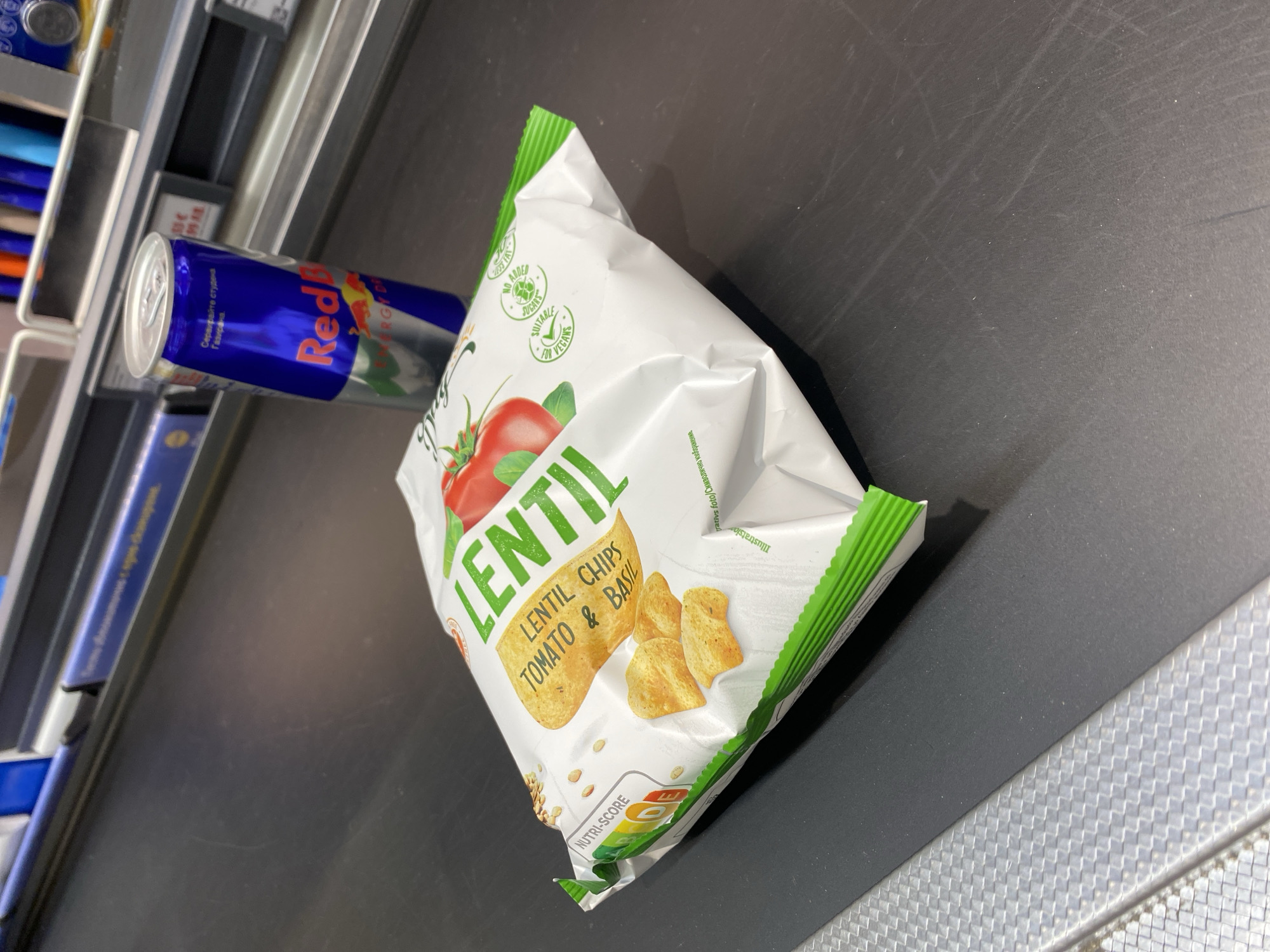 A slightly tilted, medium shot depicts a bag of "Lentil" brand lentil chips with a tomato and basil flavor, alongside a can of Red Bull energy drink, positioned on a black conveyor belt checkout counter. The chips bag is predominantly white with green accents and features images of tomatoes, basil, and lentil chips. The Red Bull can is blue and silver. The scene appears to be inside a store, with a metal shelf visible behind the Red Bull can. A section of Bulgaria is found on the Red Bull can, suggesting a likely location in Bulgaria. The photograph was taken inside a store, in the city of Plovdiv, Bulgaria.