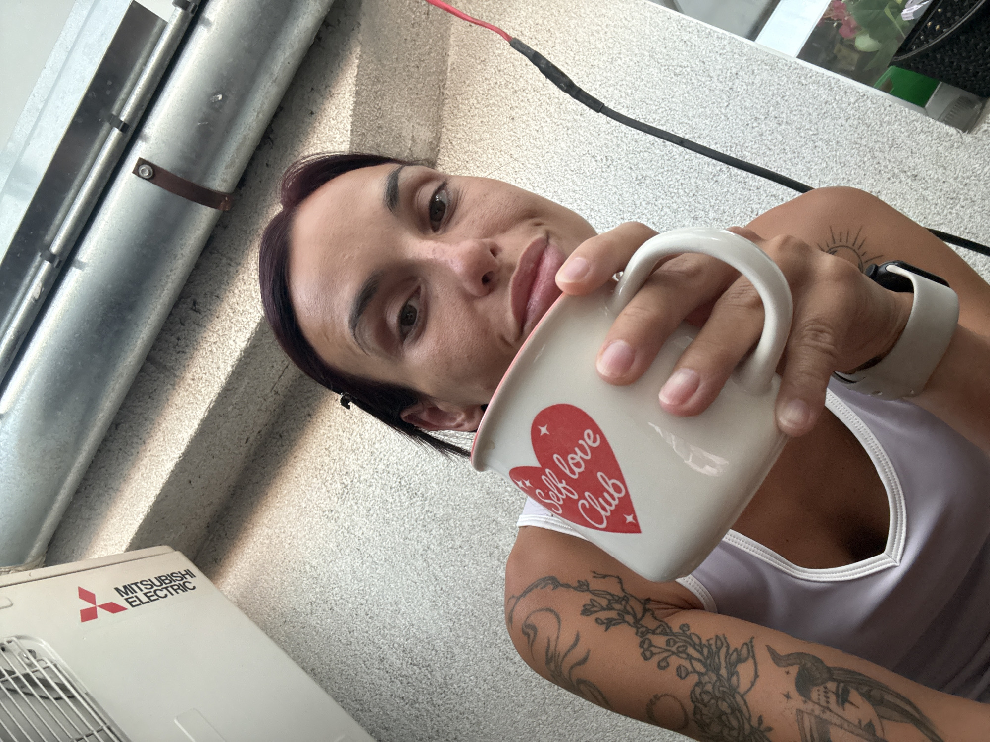 A woman is taking a selfie while holding a mug. She is wearing a white tank top and has tattoos on her arm. The mug has a red heart with the words "Self love Club" written on it. In the background, there is an air conditioning unit with the Mitsubishi Electric logo. The scene appears to be outdoors, possibly on a balcony or patio, with a metal pipe and a textured wall visible. There is a black cable with a red wire attached to it running down the wall. The image does not provide any information about the location in Pleven, Bulgaria.