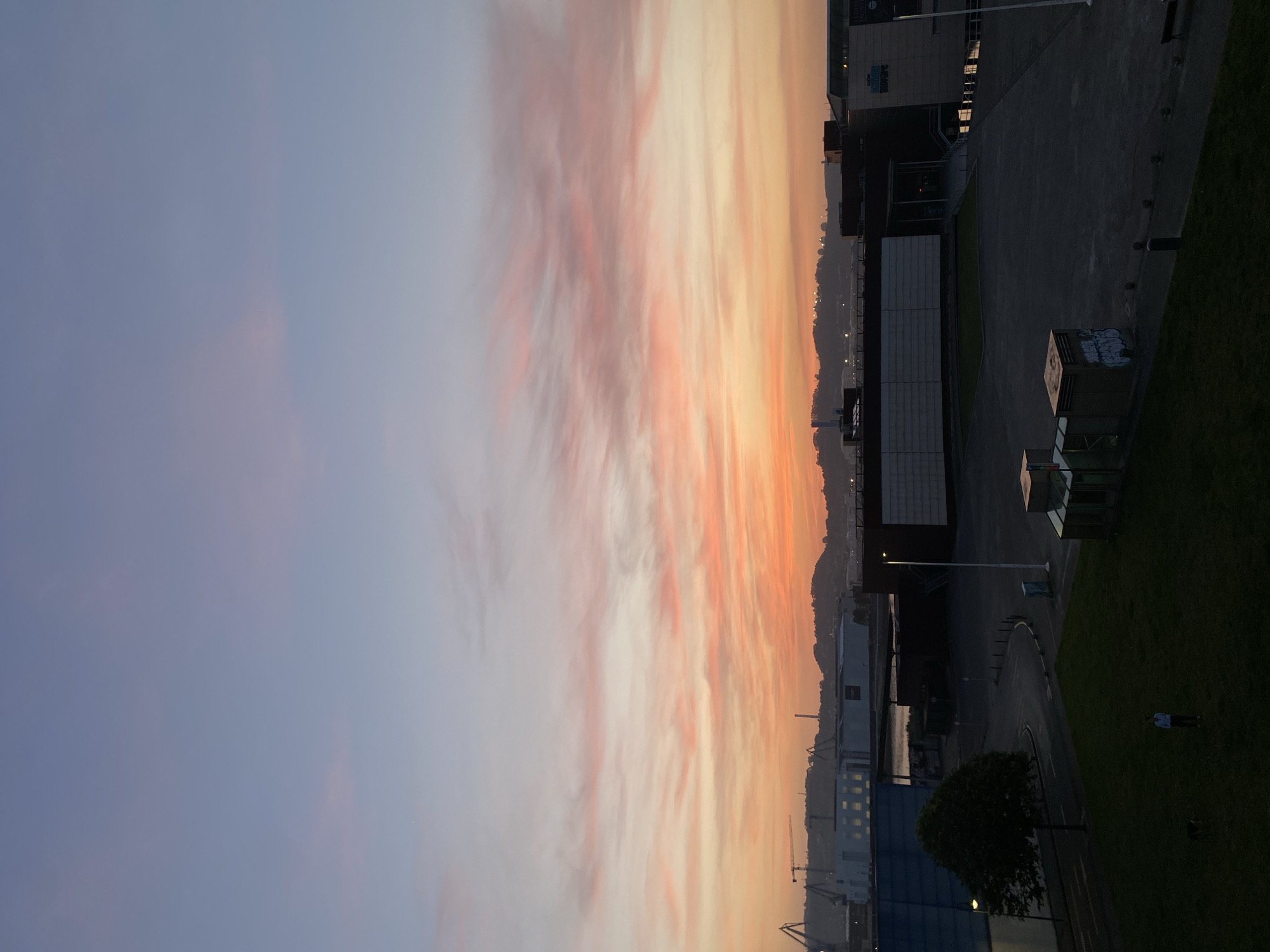 The image presents a twilight cityscape. The sky exhibits a gradient of color from a faint blue to orange. The horizon contains indistinct structures, including buildings and cranes, suggestive of a harbor or industrial zone. Adjacent to this scene lies a paved area, alongside modern architectural structures, including buildings and possibly park facilities. Adjacent to the built environment, there is green space, possibly a park or grassy area, where a person stands. The scene is in Gijón/Xixón, Spain.