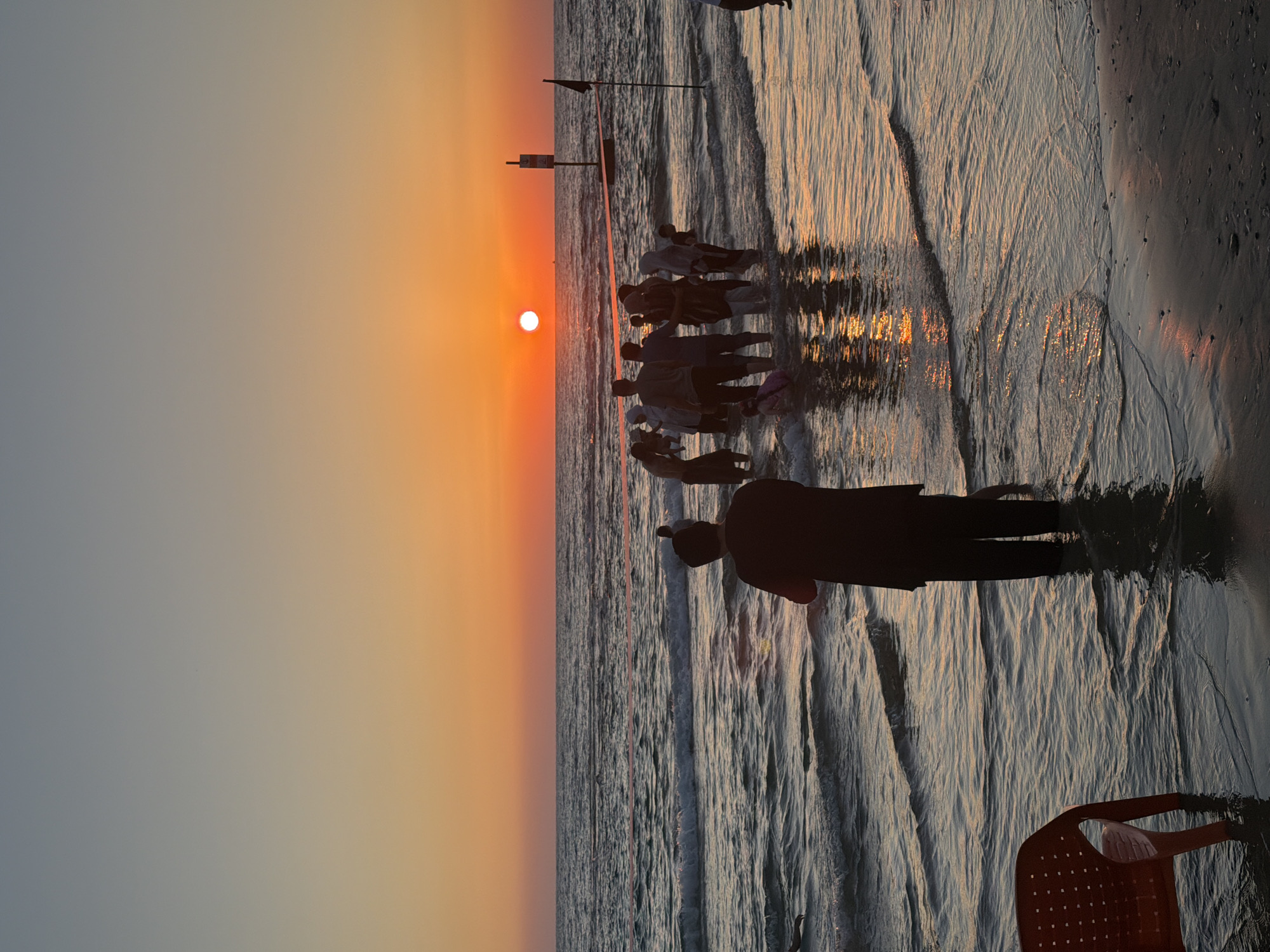 This image depicts a sunset on a beach in Tel Aviv, Israel. The sun is a bright orange disc low on the horizon, casting a warm glow across the sky and reflecting on the water. Gentle waves roll onto the shore. A group of people, some wearing headscarves, are wading in the shallow water, enjoying the sunset. In the foreground, a person in dark clothing stands facing the sea, their back to the camera. An empty orange plastic chair sits on the wet sand to the left. A red rope barrier runs horizontally across the water in the middle ground, with a black flag on a pole visible to the right. A signpost is also present in the water. The overall scene conveys a peaceful, communal atmosphere at the end of the day.