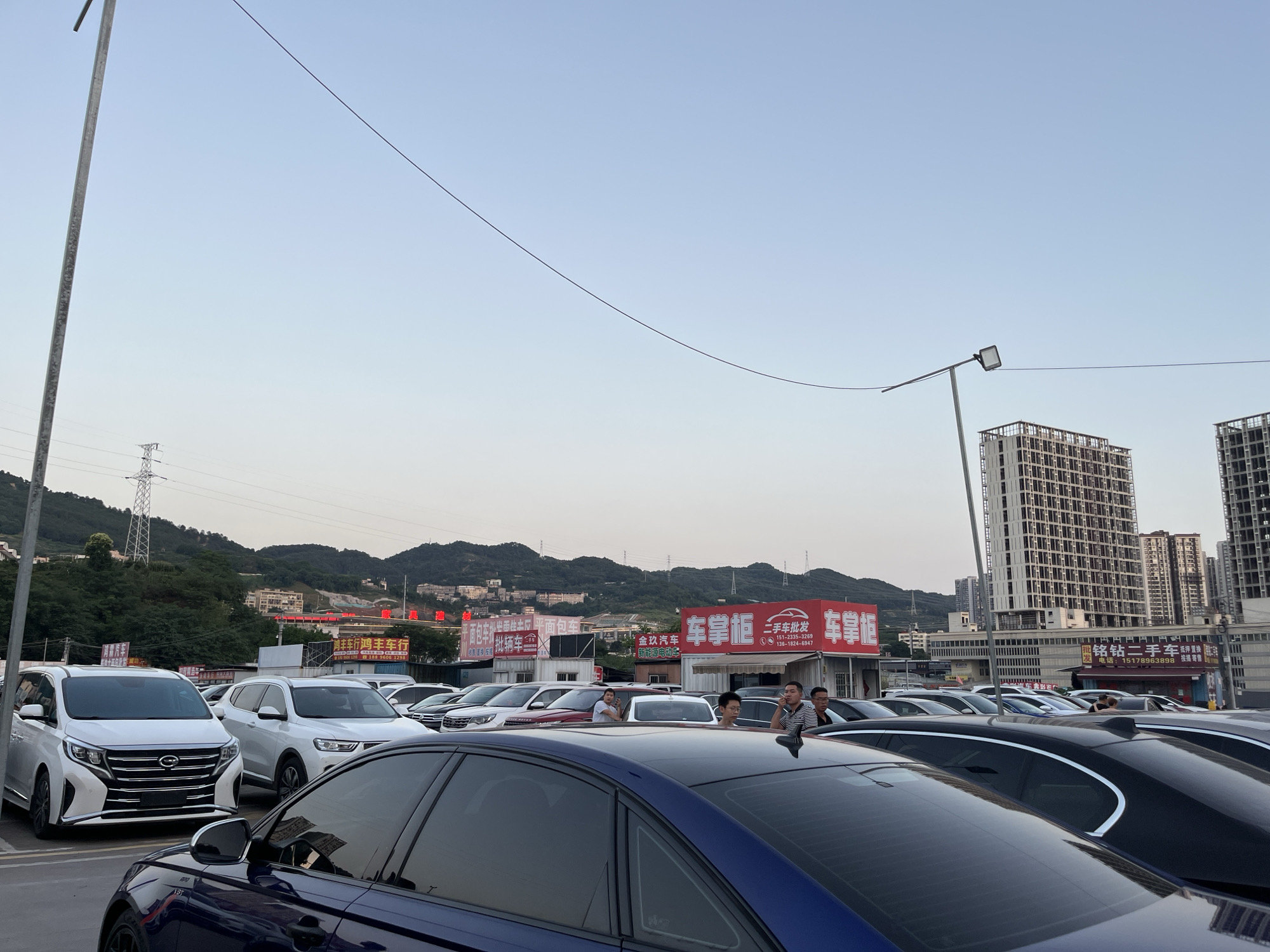 The image shows a parking lot full of cars in Banan, China. The location seems to be an area with businesses related to the automotive industry, evidenced by signs such as "二手车批发" (second-hand car wholesale) and names of car companies like "金玖汽车" (Jin Jiu Auto). The area has mountains in the background and a few buildings, some under construction. There are a few people standing among the cars.
