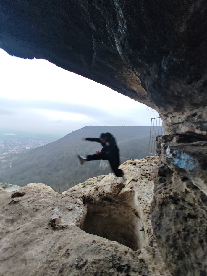 The scene is set within a natural rock formation on the Shumen Plateau, overlooking the city of Shumen, Bulgaria. A dark, textured rock overhang forms the upper portion of the frame, functioning as the ceiling of a natural shelter or cave entrance. Below, a rugged, light-colored rock platform constitutes the ground, featuring a distinct, roughly rectangular depression or hole in the lower-center.
In the mid-ground, a single individual is captured mid-air, blurred due to motion, suggesting a jump or kick action. The person is dressed in dark clothing, including a jacket and trousers, with light-colored shoes visible on their extended left leg. Their body is angled to the left, with arms outstretched. To the right of the individual, a grey metal railing or gate structure is partially visible, affixed to the rock face, which also exhibits patches of blue graffiti.
The background reveals a panoramic landscape under an overcast sky. Immediately behind the rock platform are densely forested hills. Beyond these hills, the city of Shumen is discernible in the distance, characterized by numerous buildings and an urban spread.