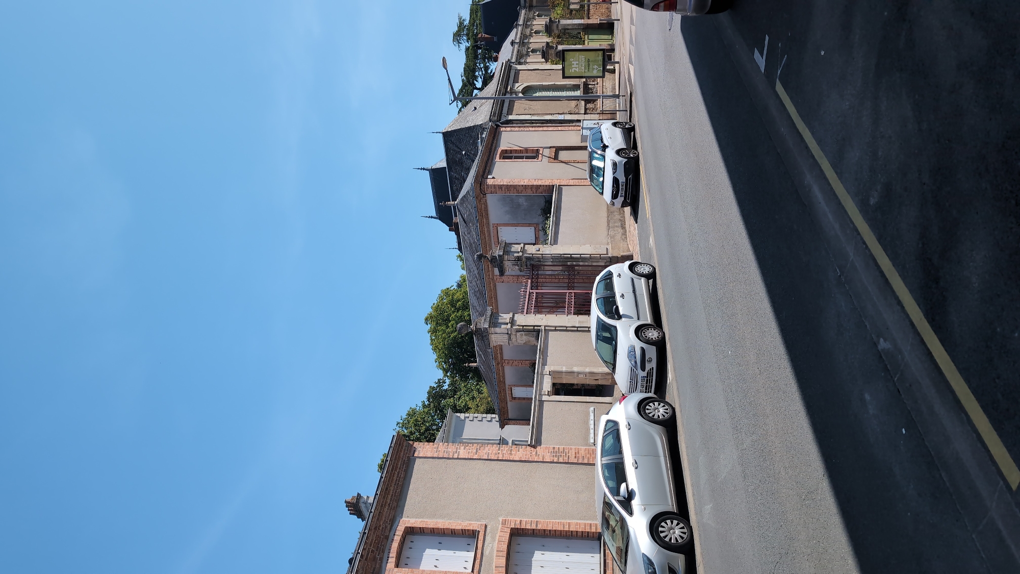 The image shows a street in Cholet, France, with several cars parked along the side of the road. In the background, there are buildings with distinctive architectural features, including a large gate with ornate ironwork. The sky is clear and blue, indicating a sunny day. The street appears to be a typical urban environment with a mix of residential and possibly commercial properties. The cars are of various makes and models, suggesting they belong to residents or visitors. There are no people visible in the image, and no particular actions or interactions are taking place. The overall scene is calm and uneventful, capturing a slice of everyday life in the city.
