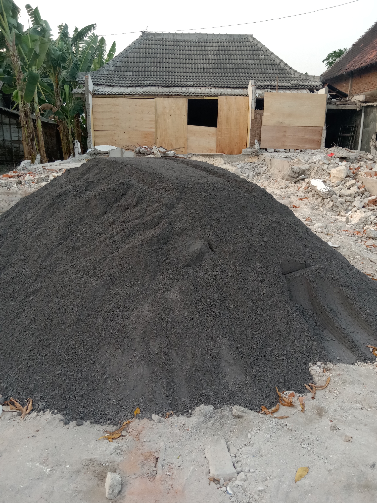 A pile of dark gray construction material, likely sand or fine gravel, dominates the foreground of this outdoor scene. The material is heaped in a large, irregular mound. Behind the pile, the remains of a building are visible, suggesting demolition or ongoing construction. Sections of a roof with gray tiles are intact, supported by wooden planks and exposed framing. The walls appear to be partially constructed with wood panels and some brickwork. Debris, including broken concrete and bricks, is scattered around the base of the structure. In the background to the left, several large banana trees with broad green leaves stand tall. The ground is a mix of dirt and scattered gravel. The sky is overcast, indicating a daytime scene. The location is described as Stiaji, Indonesia.