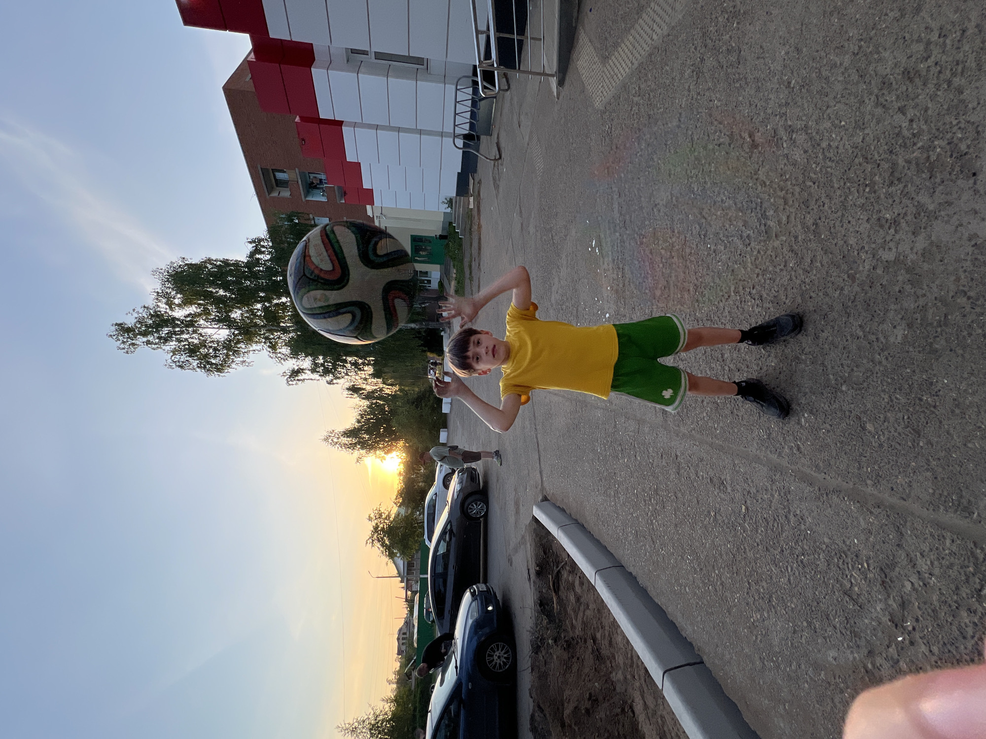 The image shows a boy in a yellow shirt and green shorts, likely playing with a soccer ball in Noviy, Russia, as the image title implies. He's standing on a paved surface, seemingly a sidewalk or parking area. A large soccer ball is positioned near his head, as if he's about to hit it. In the background are parked cars, trees, and a building with a red and white facade. The scene is illuminated by the setting sun, creating a warm glow in the sky. Another person can be seen standing near the cars in the distance, likely a spectator.