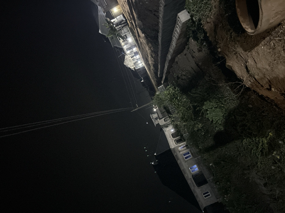 An aerial perspective captures a stark contrast between a black sky and a dimly lit urban landscape. A tall building with faint lights shining through its windows stands out against the darkness. A wall with vegetation sits to the right, and a pipe is placed along the wall's upper edge. Several power lines cut across the black sky above.