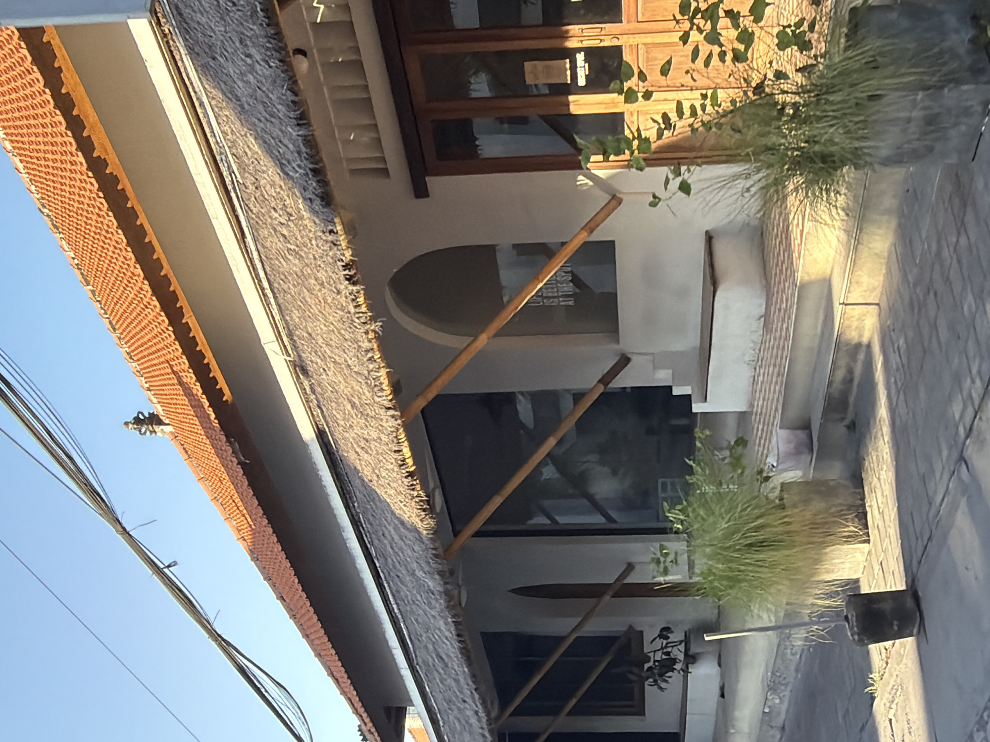 The image captures a sunlit facade of a building with a thatched roof overhang, suggesting a tropical or coastal setting. The building's exterior is white, with arched windows and wooden doors. A paved walkway leads up to the entrance, flanked by potted plants. A portion of an electrical wire is visible in the upper left quadrant of the image, indicating urban infrastructure. The time of day appears to be late afternoon, as evidenced by the long shadows cast across the scene. The overall impression is one of a welcoming, possibly commercial establishment. Based on the architectural style and the presence of lush greenery, it is plausible that this building is located in North Kuta, Indonesia, a region known for its blend of traditional and modern aesthetics and vibrant outdoor spaces.