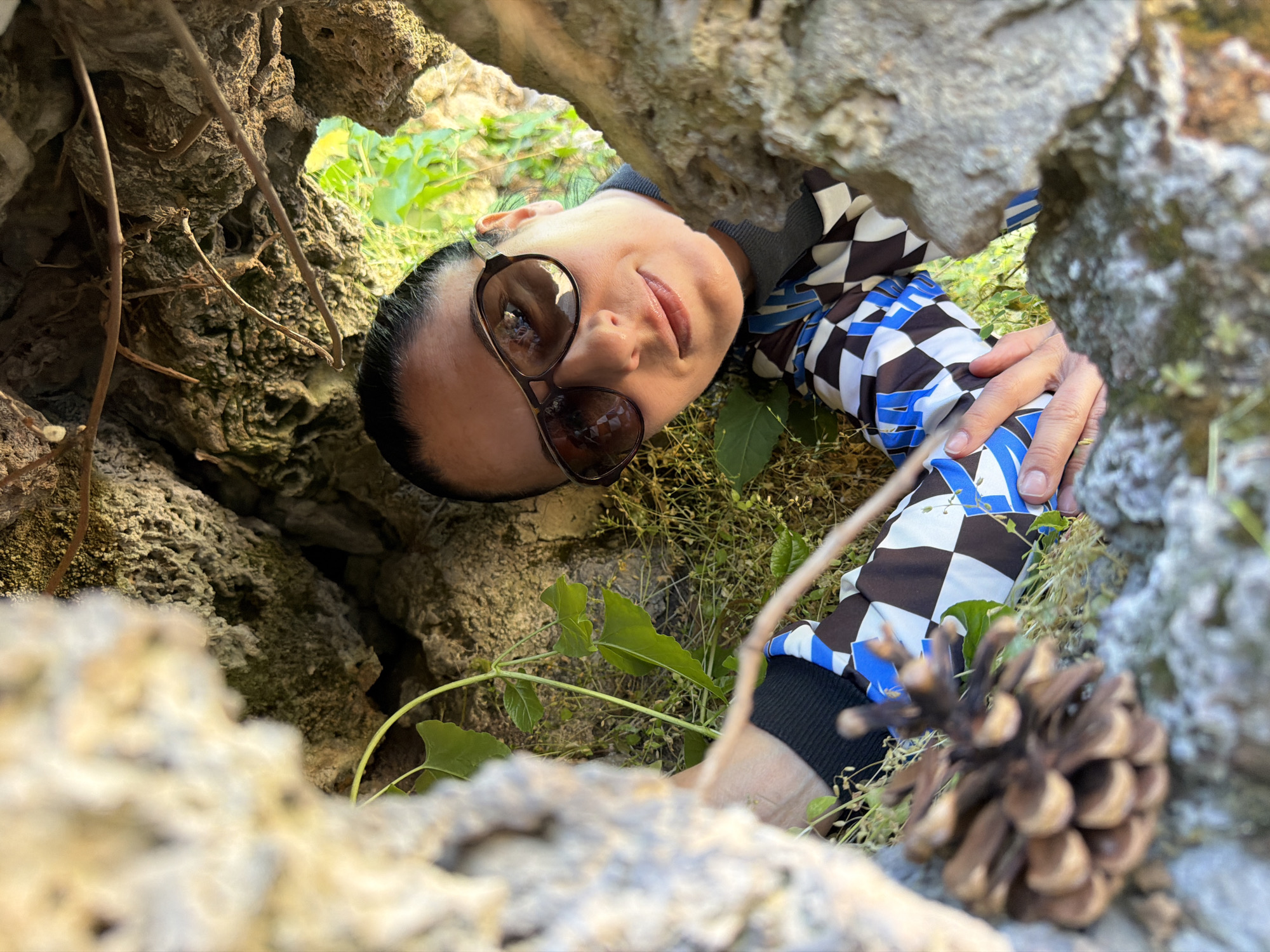 The image shows a woman in sunglasses and a checkered patterned jacket lying down, looking up through a natural opening in rocks. The woman's face is visible, and her hand is positioned near a pinecone. The surroundings consist of rocks, grass, and other vegetation. The location is Pleven, Bulgaria.