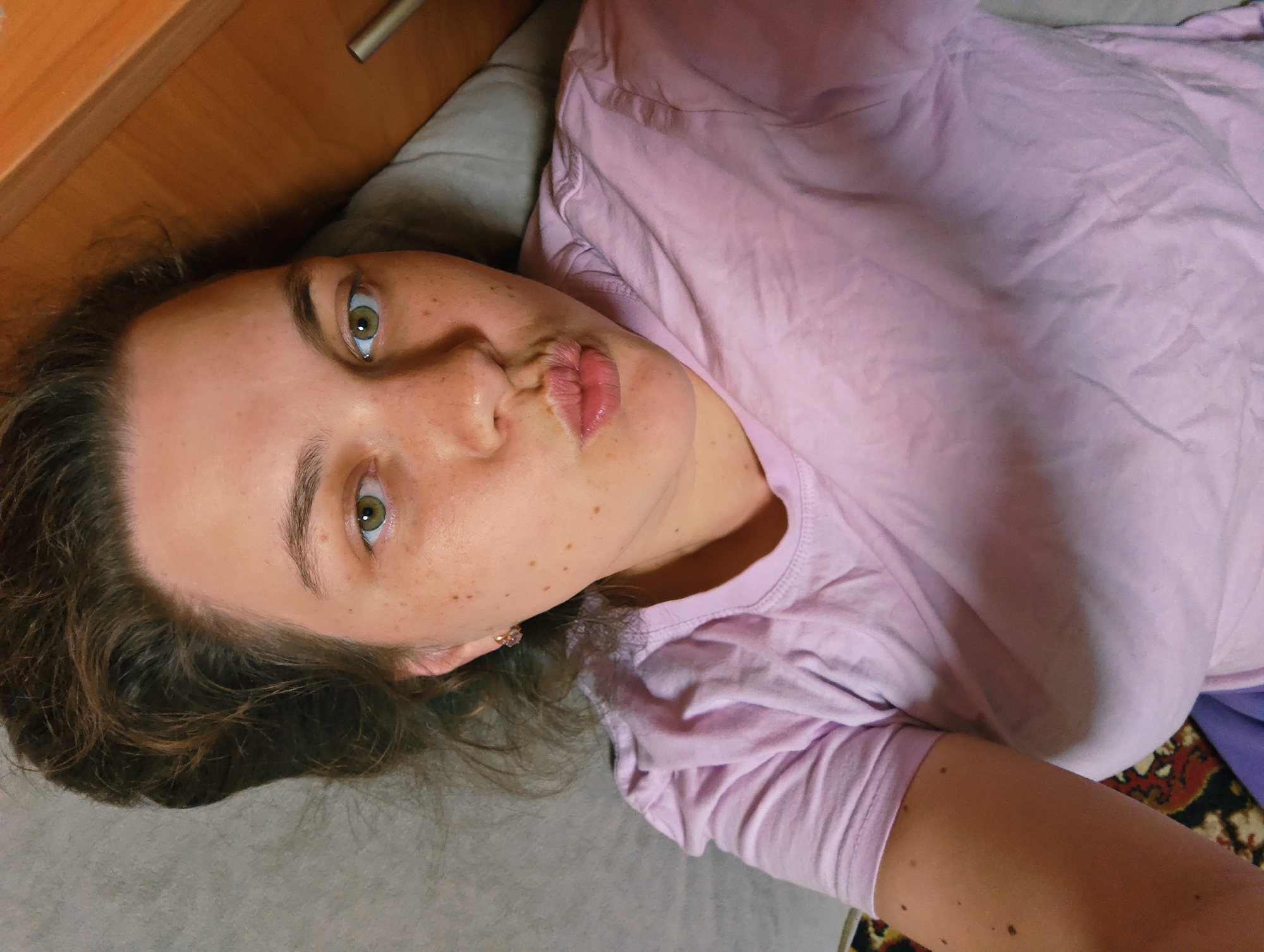 A woman is lying on her back, looking up and puckering her lips for a kiss. She is wearing a light pink t-shirt. Her hair is dark brown and curly. She has fair skin with freckles and green eyes. She is lying on a gray surface, possibly a bed or couch. A wooden cabinet or headboard is visible in the upper left corner of the frame. A rug with a dark, intricate pattern is visible in the lower right corner. The photograph may have been taken in Уральское, Russia, but no local landmarks or objects clearly identify the location.