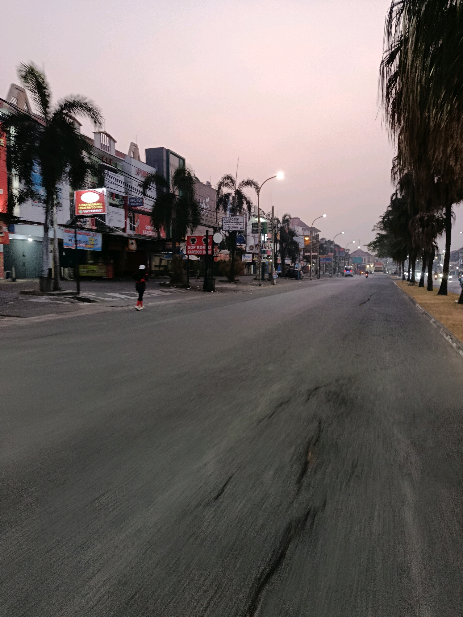 The image depicts a street scene in Bekasi, Indonesia, at dusk. The sky is a soft gradient of pink and orange, indicating either sunrise or sunset. On the left side of the road, there are buildings with shops, including an "APOTEK" (pharmacy) and signs for "YAMAHA" and "SAMSUNG". Palm trees line the street, with some in the foreground obscuring parts of the buildings. A person wearing dark clothing with a white head covering and orange shoes is visible in the middle of the road, seemingly engaged in an activity such as rollerblading or skateboarding, given the posture and the presence of what appear to be roller skates or similar footwear. The road itself is paved and shows signs of wear, with visible cracks and textured patterns due to motion blur, suggesting the image was taken from a moving vehicle or device. Further down the street, streetlights are illuminated, and the headlights of approaching vehicles are visible, indicating continued activity despite the low light conditions. The right side of the road features a sidewalk and more palm trees. The overall impression is of a semi-urban environment transitioning from day to night.