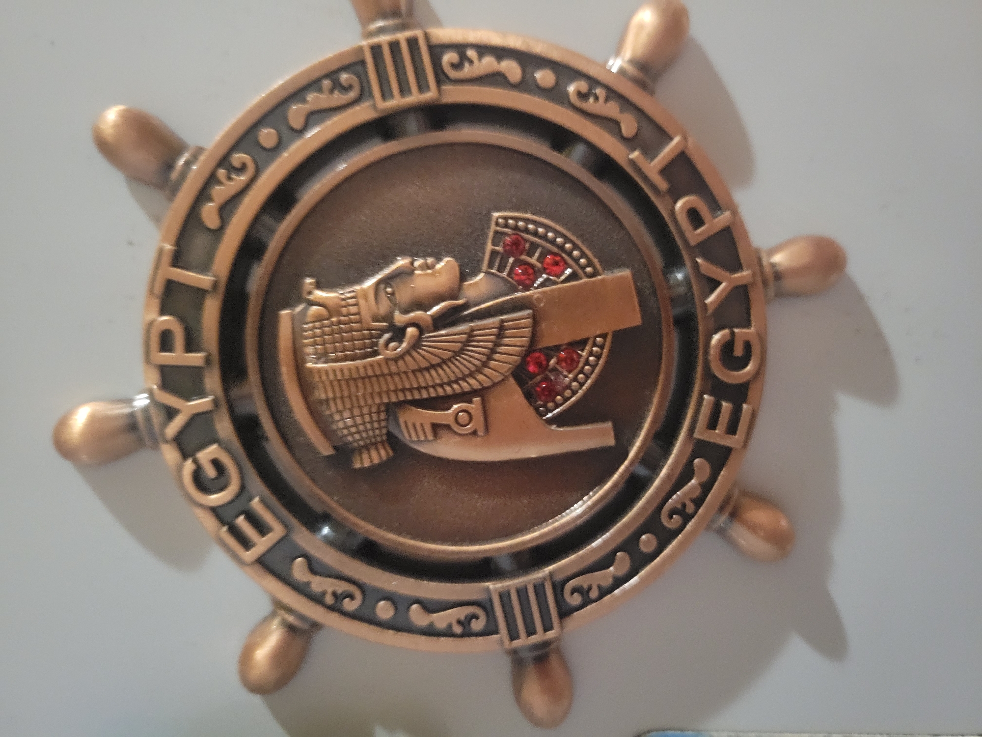 The image shows a close-up, angled shot of a decorative, circular emblem with a nautical wheel design. The emblem features a profile of an Egyptian pharaoh's head in the center, adorned with a headdress and necklace, the latter decorated with red stones. The word "EGYPT" is embossed in capital letters around the outer rim of the emblem, separated by decorative elements. The emblem appears to be made of a metallic material with a bronze finish. The background is a plain, light-colored surface, likely a wall or a door, and is out of focus. The lighting creates soft shadows around the emblem. The image does not contain any discernible landmarks or contextual information that would allow for the determination of its location in Belaazyorsk, Belarus, or any other city.