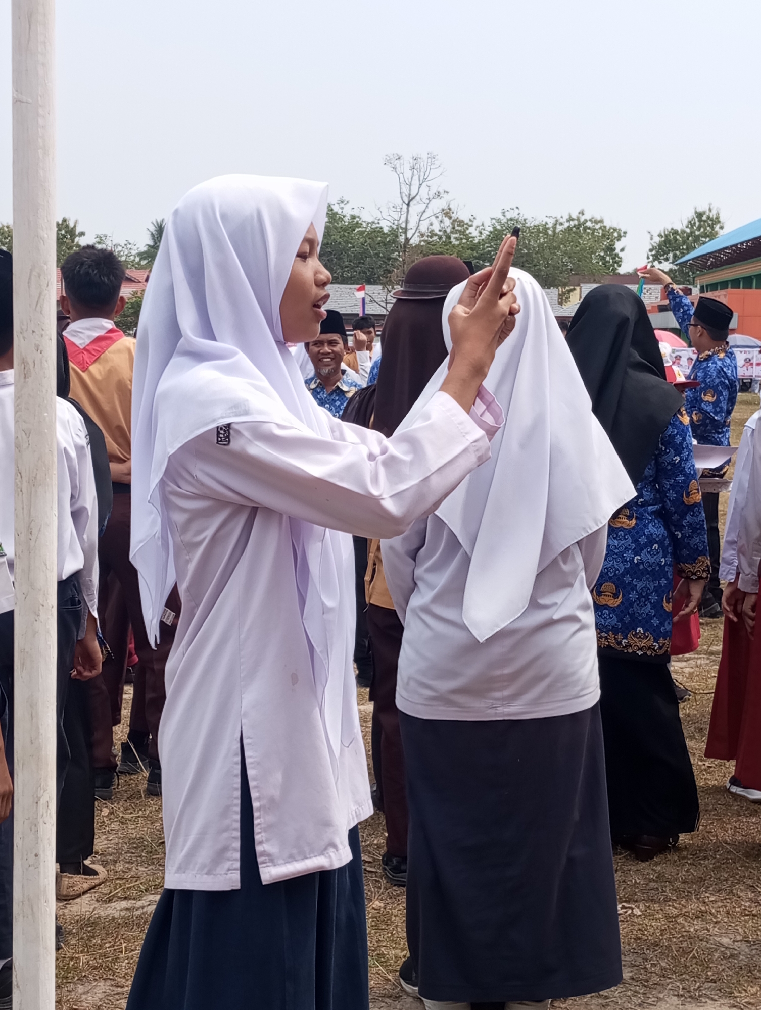 A group of people, mostly young women in white hijabs and dark skirts, are gathered outdoors. One young woman in a white hijab and a light purple shirt is the most prominent figure, pointing her index finger upwards and to the right, seemingly addressing someone. She is wearing a dark blue skirt and her hair is partially visible under her hijab. Behind her, other individuals are visible, some in similar attire, and one man in the background is smiling broadly. The environment appears to be a grassy field with some trees and buildings in the distance. The sky is overcast. The scene suggests an outdoor event or gathering in Pematang Reba, Indonesia.