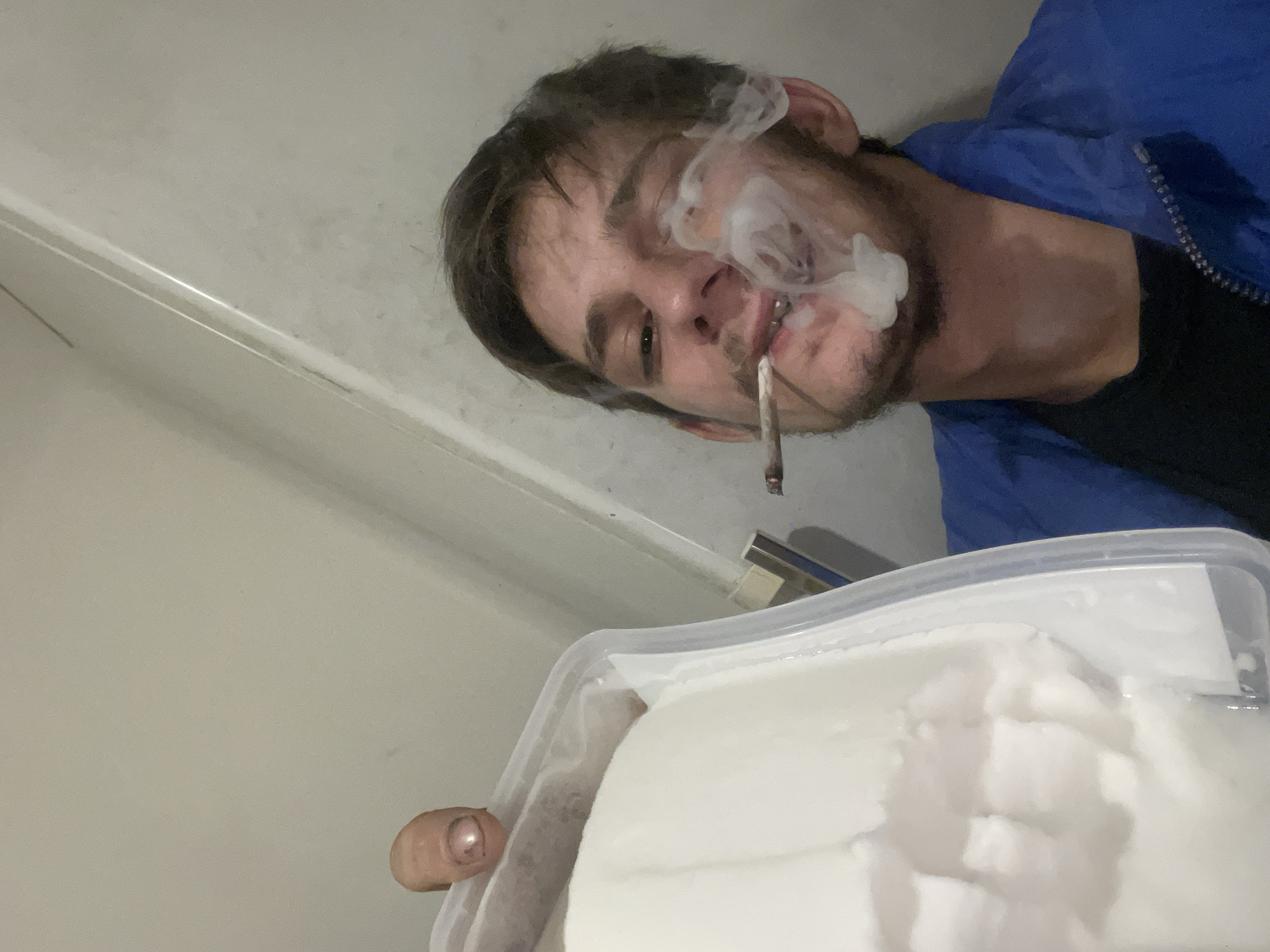 A man is holding a large container of white substance while smoking a rolled cigarette. The man has short, dark hair and a thin mustache. He is wearing a blue jacket and a black shirt underneath. Smoke is visible coming from his mouth. The location appears to be indoors, with a light-colored wall and door visible in the background. The exact location within the city of Unknown, Unknown, cannot be determined from the image.