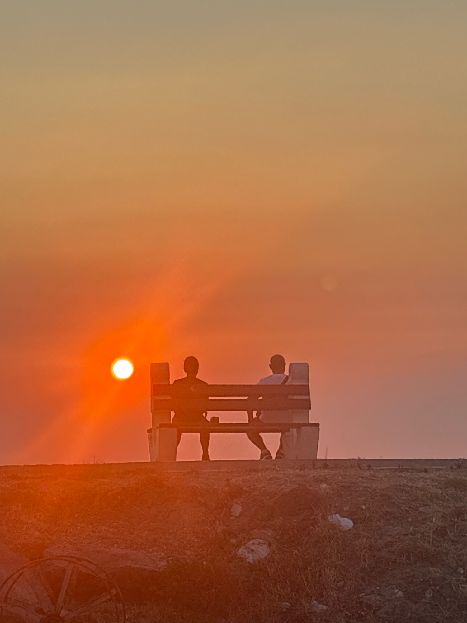 The image depicts a sunset scene in Bar, Montenegro. Two individuals are silhouetted on a park bench, facing the setting sun. The sun, a bright orange orb, is low on the horizon, casting a warm glow across the sky, which transitions from a pale yellow at the top to a vibrant orange near the horizon. The ground in the foreground is covered with grass and sparse vegetation, with some debris visible. A portion of an old wooden wheel is present in the lower left corner. The bench appears to be made of wood and concrete. The overall atmosphere is serene, with the focus on the natural beauty of the sunset and the quiet contemplation of the two figures.