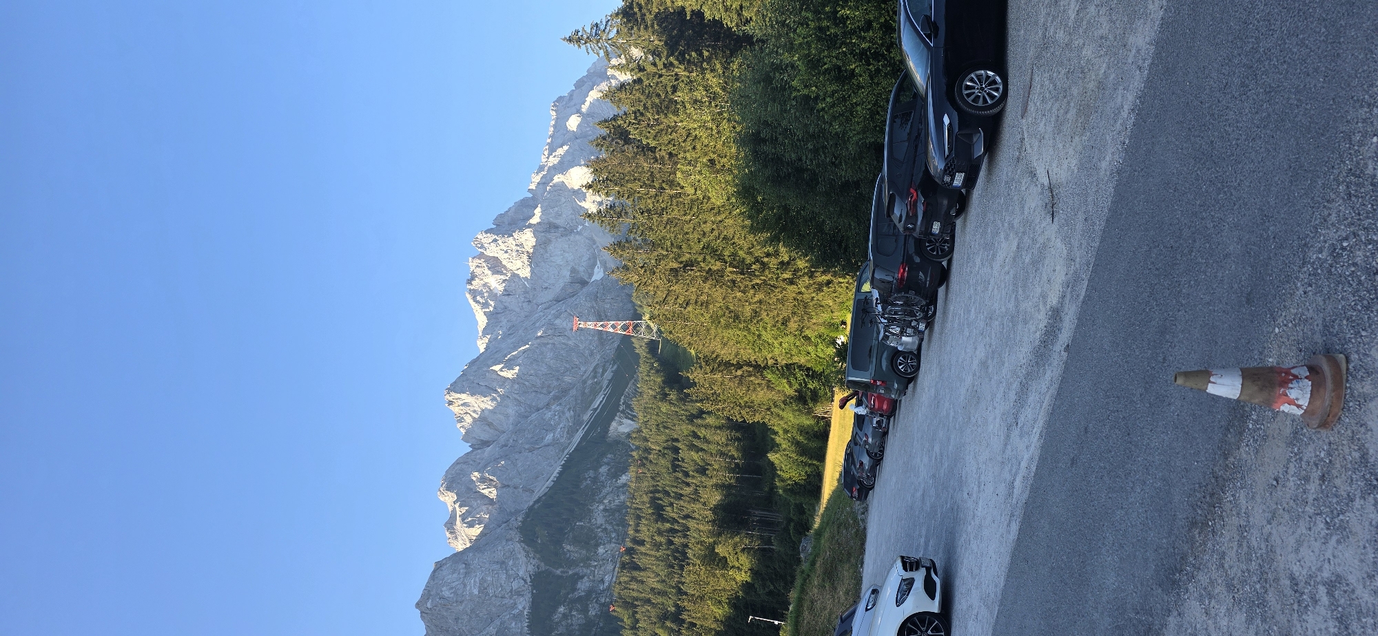 The image shows a parking lot in Grainau, Germany, with the Zugspitze mountains in the background. Several cars are parked in the lot, and a red and white tower is visible on the mountainside. The sky is clear and blue. In the foreground, a traffic cone is placed on the ground. The scene appears to be a parking area for tourists visiting the Zugspitze region.