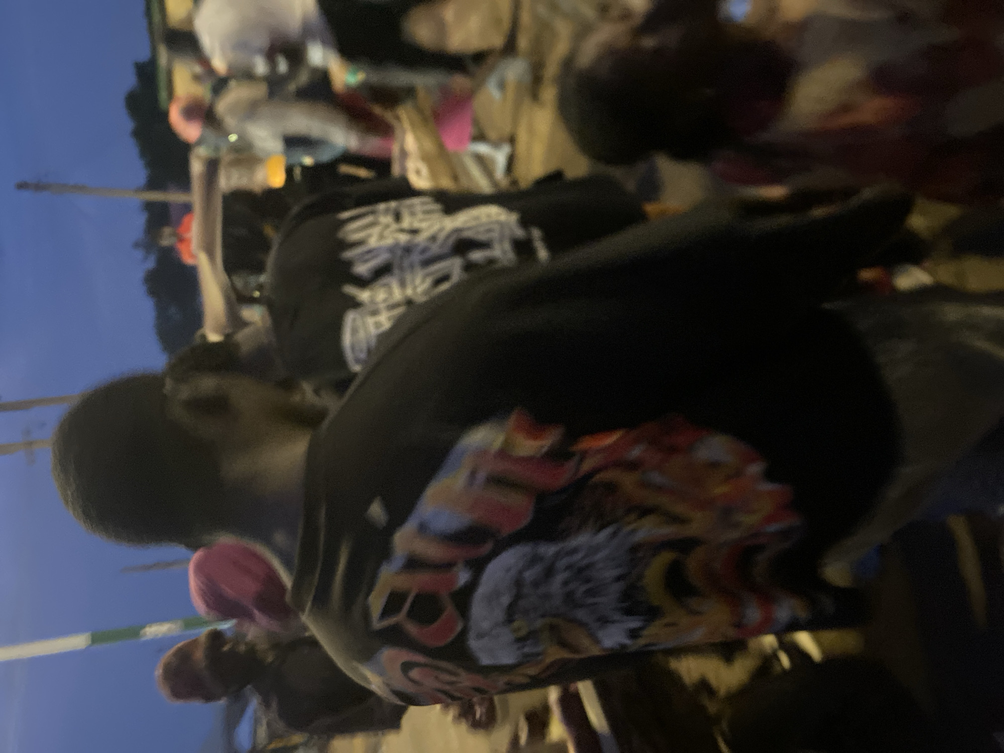 The image depicts a group of people in an outdoor urban setting, likely in Bukuru, Nigeria, as suggested by the prompt. The lighting indicates it is either dusk or dawn.
In the foreground, a person wearing a black long-sleeved shirt with a large graphic design on the back is the primary subject. The graphic features an eagle's head with fiery elements and text, partially obscured. The person's head is turned away from the camera.
Behind this individual, other people are visible, some with their backs to the camera. One person is wearing a pink head covering, and another is wearing a red head covering. To the right, a child wearing a pink and white garment is partially visible. Several other figures are indistinctly seen in the background, along with what appears to be a vehicle or structure.
The environment suggests a public space with some form of shelter or stall visible. The ground appears to be paved or dusty. The overall impression is of a casual gathering or street scene.