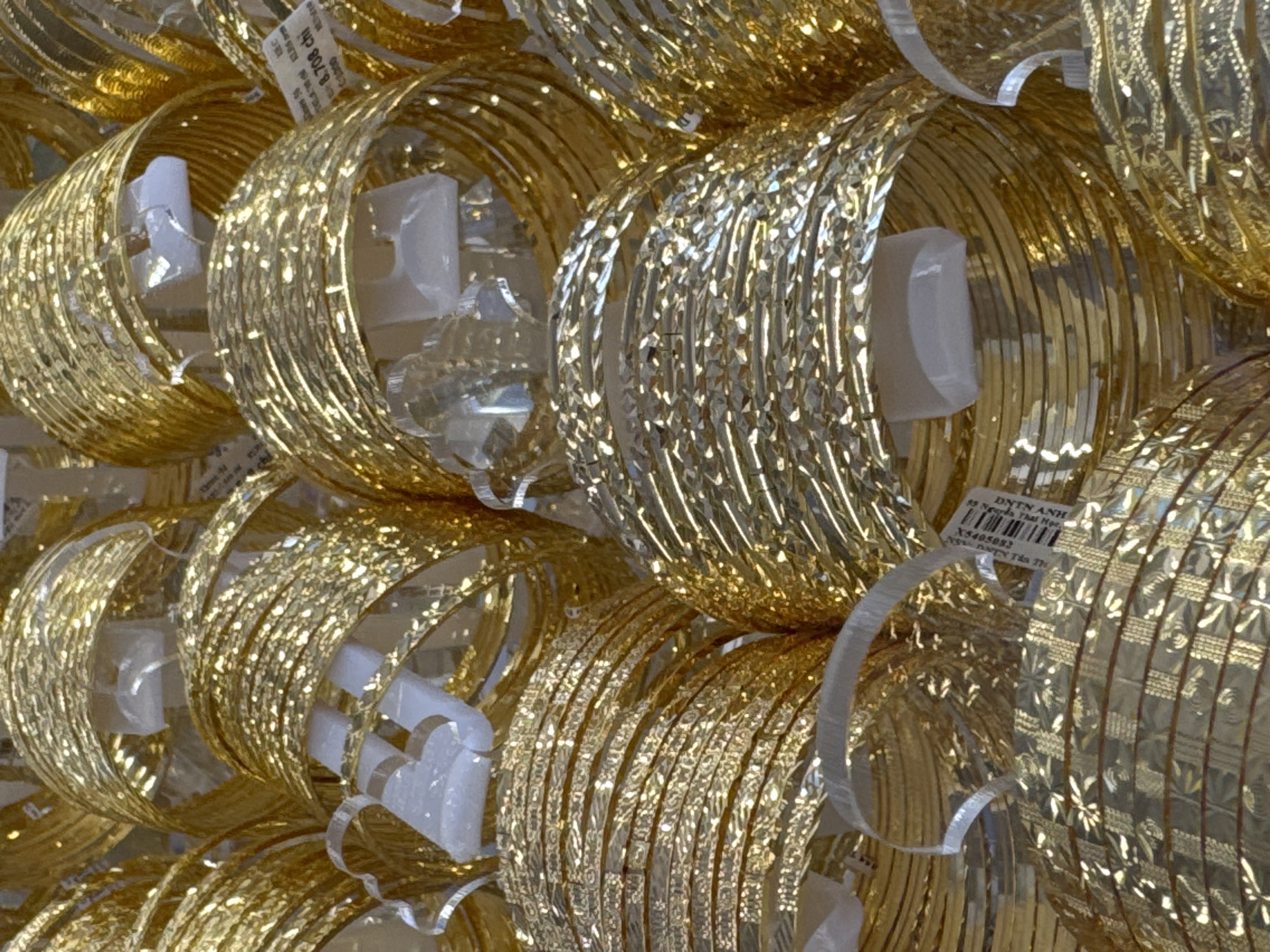 This image shows a close-up view of a display of numerous gold bangles. The bangles are stacked and arranged in a visually dense composition, with their metallic sheen reflecting light. Many of the bangles appear to be intricately patterned, with some featuring a textured, almost diamond-cut surface, and others having a smooth, ribbed appearance. Small white plastic holders are visible between some of the bangles, suggesting they are part of a retail display.
In the upper right corner, a partial label is visible with some text and a barcode. The visible text on the label includes "DNTN ANH," "99 Ngarda Thal Mor," and a product code "X5405082." The reference to "Nha Trang Ward, Vietnam" in the location context suggests this is likely a jewelry store or market stall in that region. The lighting appears to be artificial, typical of an indoor retail environment, and the overall impression is one of abundance and the richness of the gold jewelry. There are no people visible in the image, and the focus is entirely on the collection of bangles.