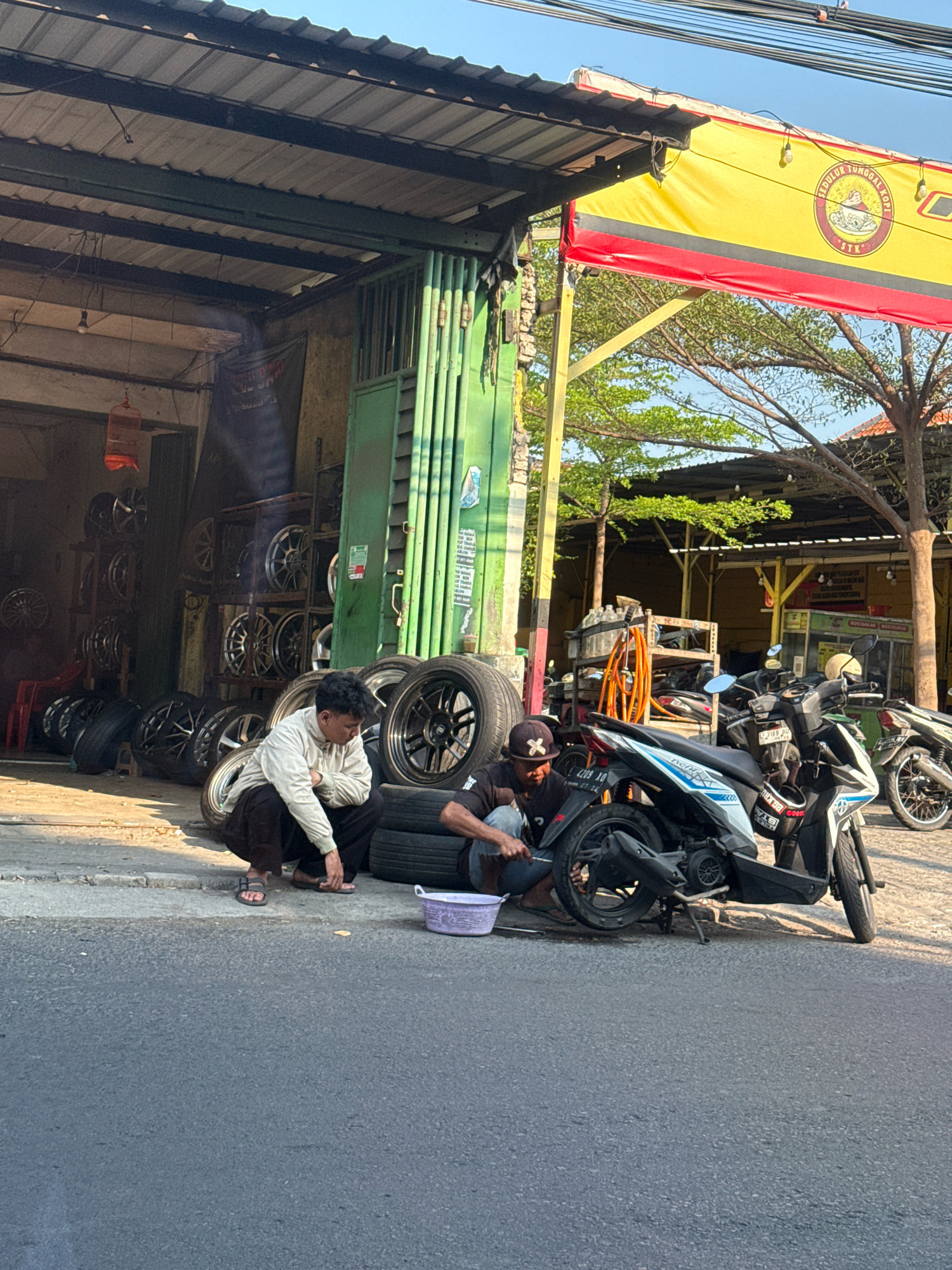 Two men are working on tires and motorcycles in front of a shop in Surabaya, Indonesia. The shop appears to be a tire repair or sales business, as there are many tires displayed on shelves and stacked on the ground. In the foreground, one man wearing a white long-sleeved shirt and dark pants is crouching next to a pile of tires. The other man, wearing a black t-shirt and a baseball cap, is kneeling beside a motorcycle, appearing to work on its tire. Several motorcycles are parked in front of the shop, with one actively being worked on. A bright yellow banner with red trim hangs above the shop, featuring a circular logo. The scene is set on a street with asphalt pavement, and the overall lighting suggests it is daytime. The environment includes a covered storefront with green metal doors and visible structural beams, along with some trees and buildings in the background.