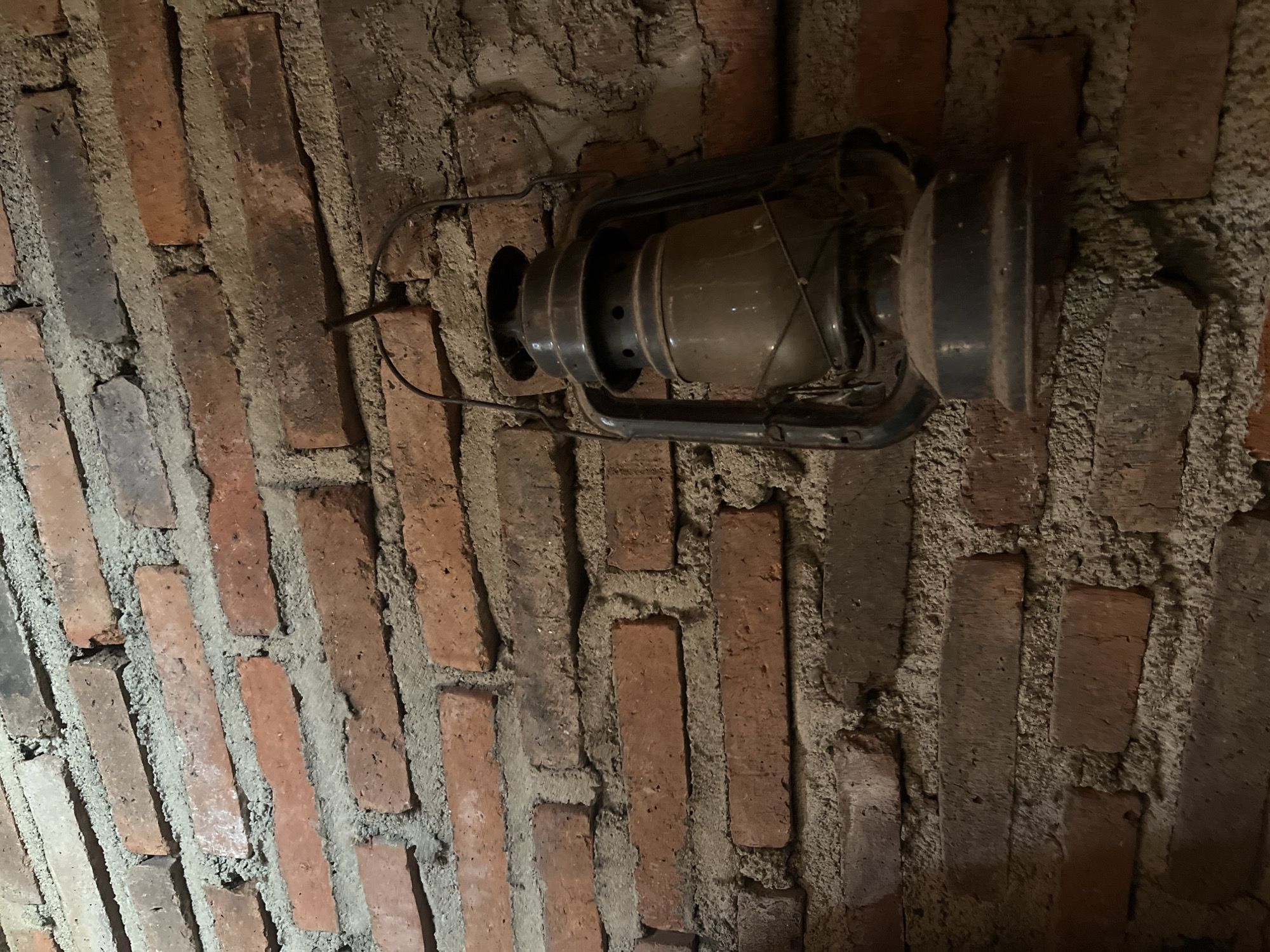 The image depicts a vintage kerosene lantern hanging on a brick wall. The wall is constructed of reddish-brown bricks of varying sizes, with visible mortar between them. The bricks appear aged and somewhat weathered. The lantern itself is dark metal, with a glass reservoir and a handle for carrying or hanging. It is positioned to the right of the frame, casting a slight shadow on the wall. The lighting suggests an indoor setting, possibly dimly lit. The scene is static and focuses on the textures of the brick and the details of the lantern. The location is identified as Glagah Kulon, Indonesia. There are no people or animals present in the image, and no actions are taking place.