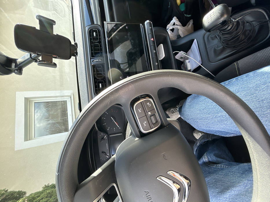 The image displays the interior of a stationary vehicle in Veauchette, France, viewed from the driver's perspective under bright daylight. A person's lower body, wearing blue denim jeans and gray sneakers, occupies the driver's seat. A black multi-function steering wheel with a silver Citroën emblem is prominent in the foreground. Behind the steering wheel, a vehicle instrument cluster with gauges is partially visible. Centrally located on the dashboard is a large, glossy infotainment screen. A black mobile phone mount is affixed to the windshield above the dashboard. To the right and lower in the frame, a six-speed manual gear stick is visible. A storage compartment below the infotainment screen contains a white power bank and a white cable. Through the vehicle's windshield, an exterior light-colored building wall with a white-framed window is visible, reflecting trees and the sky.