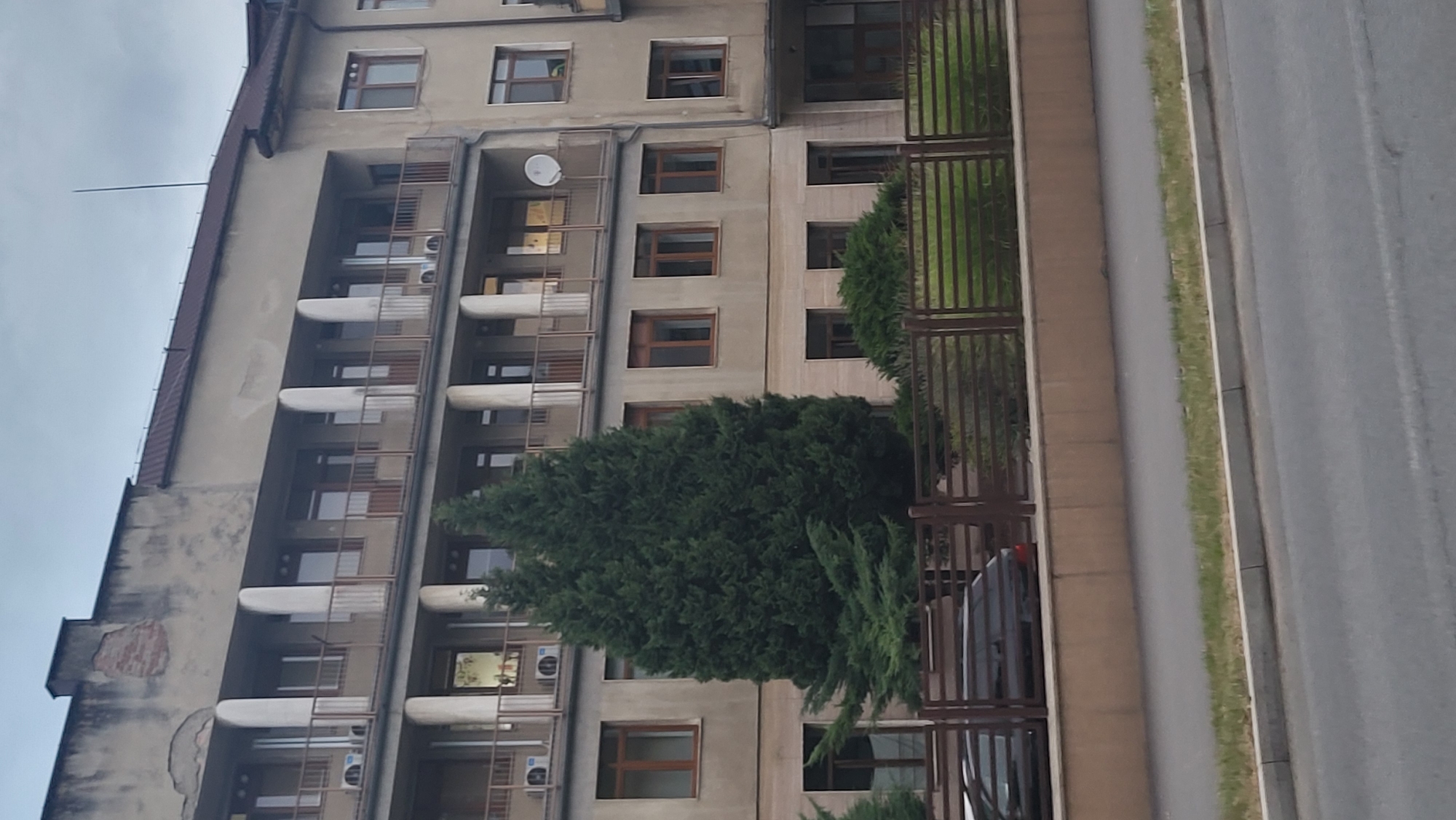 The image depicts the exterior of a multi-story building in Prešov, Slovakia. The building appears to be made of concrete and has a dated architectural style. The facade shows signs of wear and tear, with visible patches of exposed brickwork at the top. There are multiple balconies with railings, and several windows with wooden frames. Two large evergreen trees are prominent in front of the building. A dark-colored car is partially visible behind a fence. The foreground shows a section of a road and a sidewalk, with grass growing along the edge. The sky is overcast.