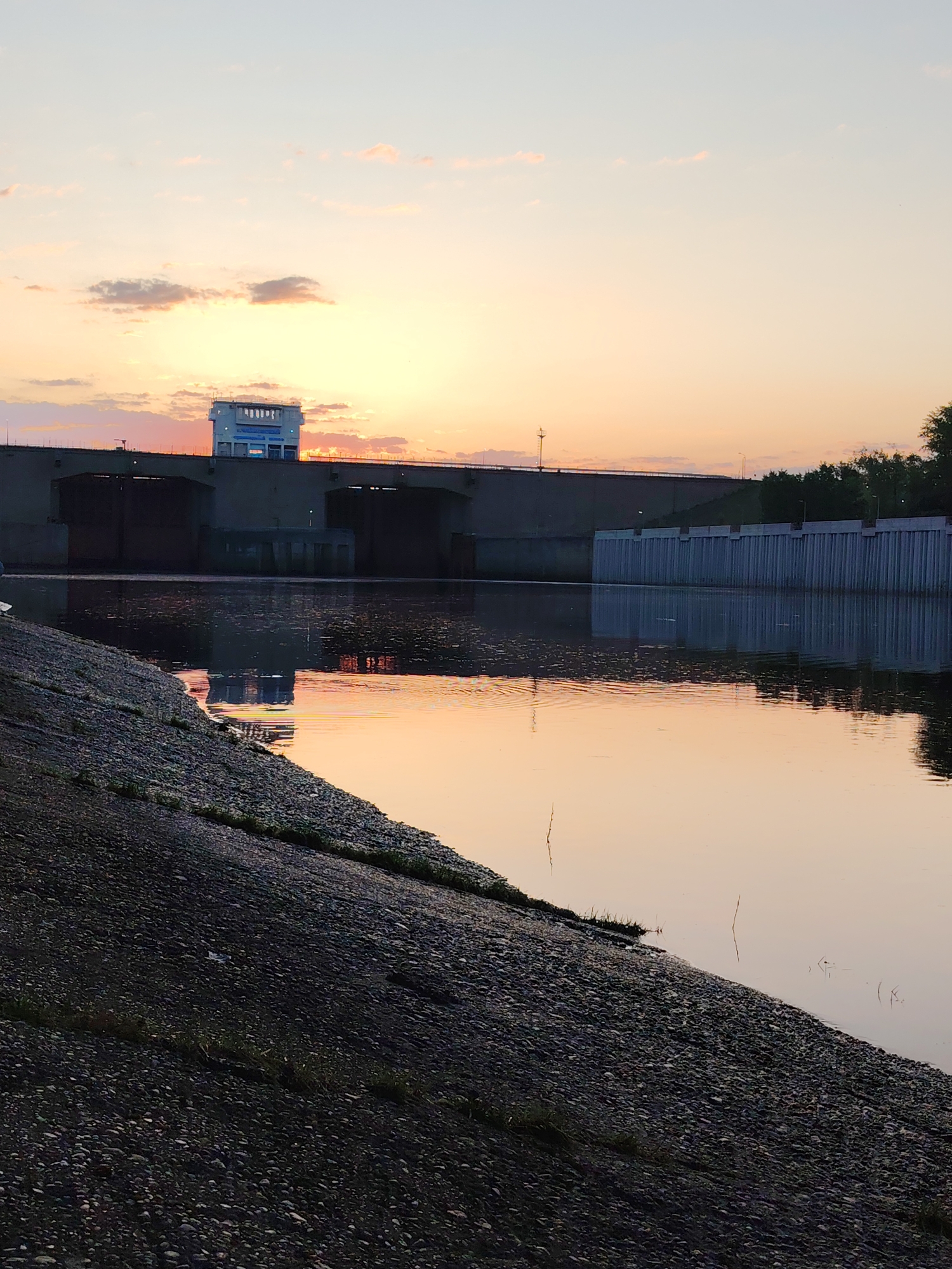 A river landscape featuring a dam and embankment at sunset. The sky shows pastel hues of orange, pink, and blue. The dam structure is concrete with rectangular sections and a prominent central building. The water reflects the colors of the sky and the structure of the dam. The embankment in the foreground is composed of pebbles and small patches of grass. The photograph appears to have been taken in or near Chaikovsky, Russia, based on the presence of a dam resembling the Chaikovsky Dam on the Kama River.
