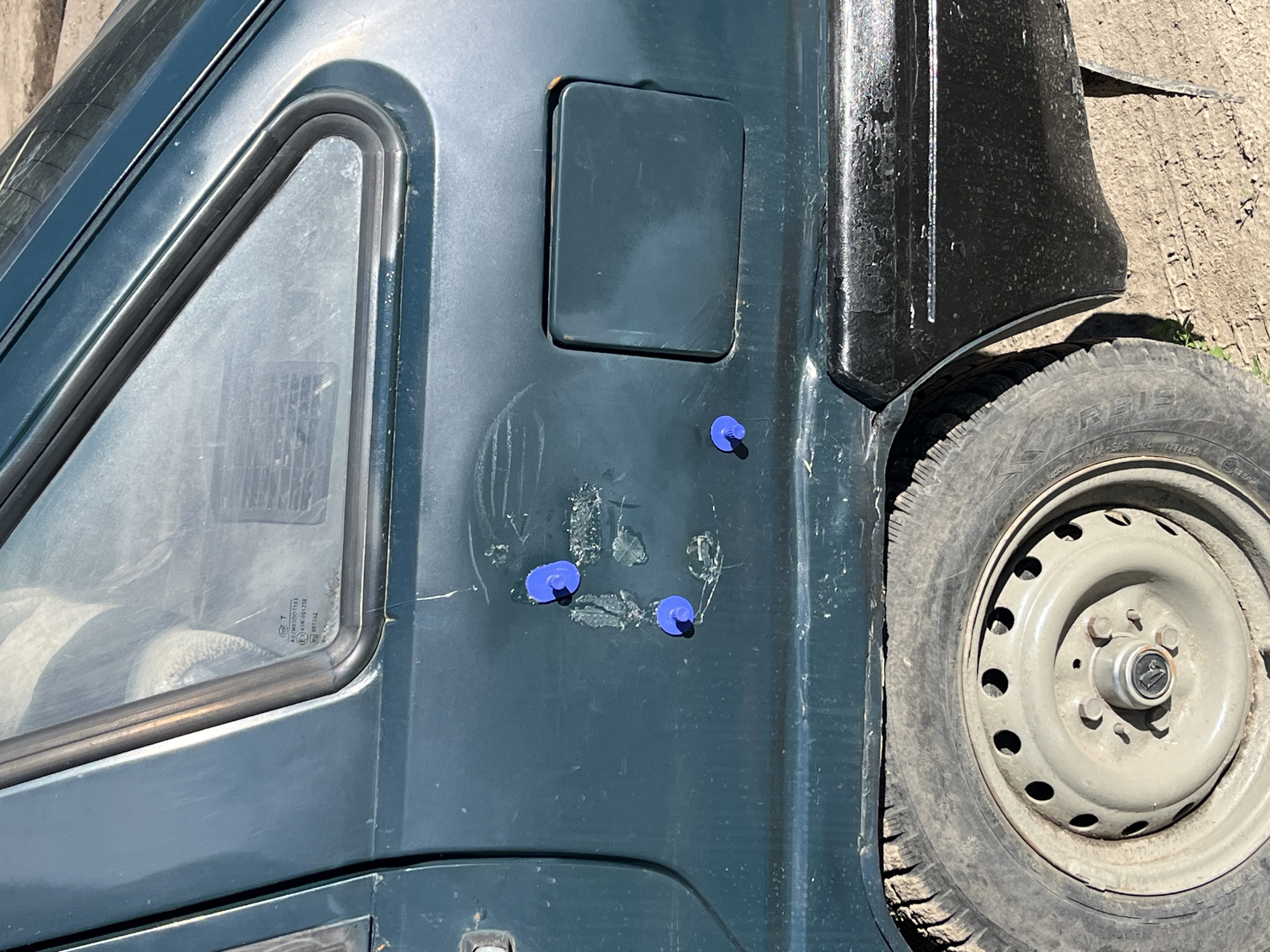 This media file is a close-up, slightly angled shot of the side of a dark green vehicle, likely a utility vehicle or small truck. The focus is on the body of the vehicle, particularly the driver's side door and fender area, with a wheel visible on the right side.
The vehicle's paint is somewhat scratched and worn, showing signs of age and use. A triangular window, outlined with black rubber trim, is prominent on the left side of the frame. The glass of the window is slightly opaque, obscuring a clear view of the interior, though a hint of a seat or fabric can be discerned. Just above and to the right of the window, a rectangular, recessed panel is visible on the vehicle's body.
Several small, bright blue, circular objects are attached to the dark green surface of the door. These appear to be some sort of caps or plugs. The background consists of rough, sandy or dirt ground with some dry vegetation.
The tire and wheel are a significant feature. The tire is a dark, used tire, and the wheel is a plain, light gray steel rim with visible lug nuts and a center cap bearing a logo. The wheel appears to be resting on the ground. The overall impression is that the vehicle is parked outdoors in a rustic or unkempt environment.
There are no people visible in the media file. The lighting suggests it is daytime, with sunlight casting shadows and highlighting the textures and imperfections of the vehicle and its surroundings. The weather appears to be dry.
Visible text is present on the glass of the triangular window, appearing as small white lettering, likely indicating safety standards or manufacturer information for the glass. One section of text is legible: "E43R001132".