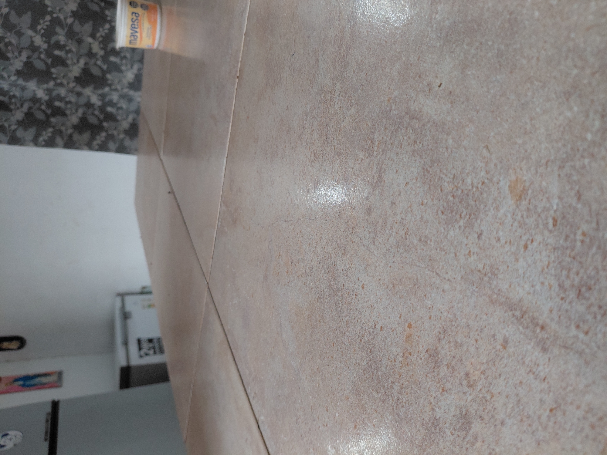 This image captures an indoor domestic scene, likely a kitchen or utility area, in Santa Ana, Venezuela. The foreground is dominated by a light beige or off-white tiled surface, possibly a floor, extending from the bottom right towards the upper left. The tiles have a mottled, slightly textured appearance with scattered darker brown and orange specks, and show reflections suggesting a polished finish or overhead lighting. A visible grout line separates the tiles.
In the midground, towards the left, a white and orange/yellow container labeled "navesa" is visible, resting on a surface. Behind this, a wall is covered with dark, patterned wallpaper or tiles featuring a floral or leafy design in shades of grey and black.
The background reveals an interior room with white walls. A large, white appliance, possibly a refrigerator or freezer, is visible on the left side of the frame, adorned with some dark markings or stickers. Below it, a dark surface, possibly a piece of furniture, is partially visible. A blurry, framed picture or poster with colorful elements (blue, pink) hangs on the wall further into the room.
No people are visible, and no specific activity is taking place; the scene is static. The lighting suggests it is daytime.