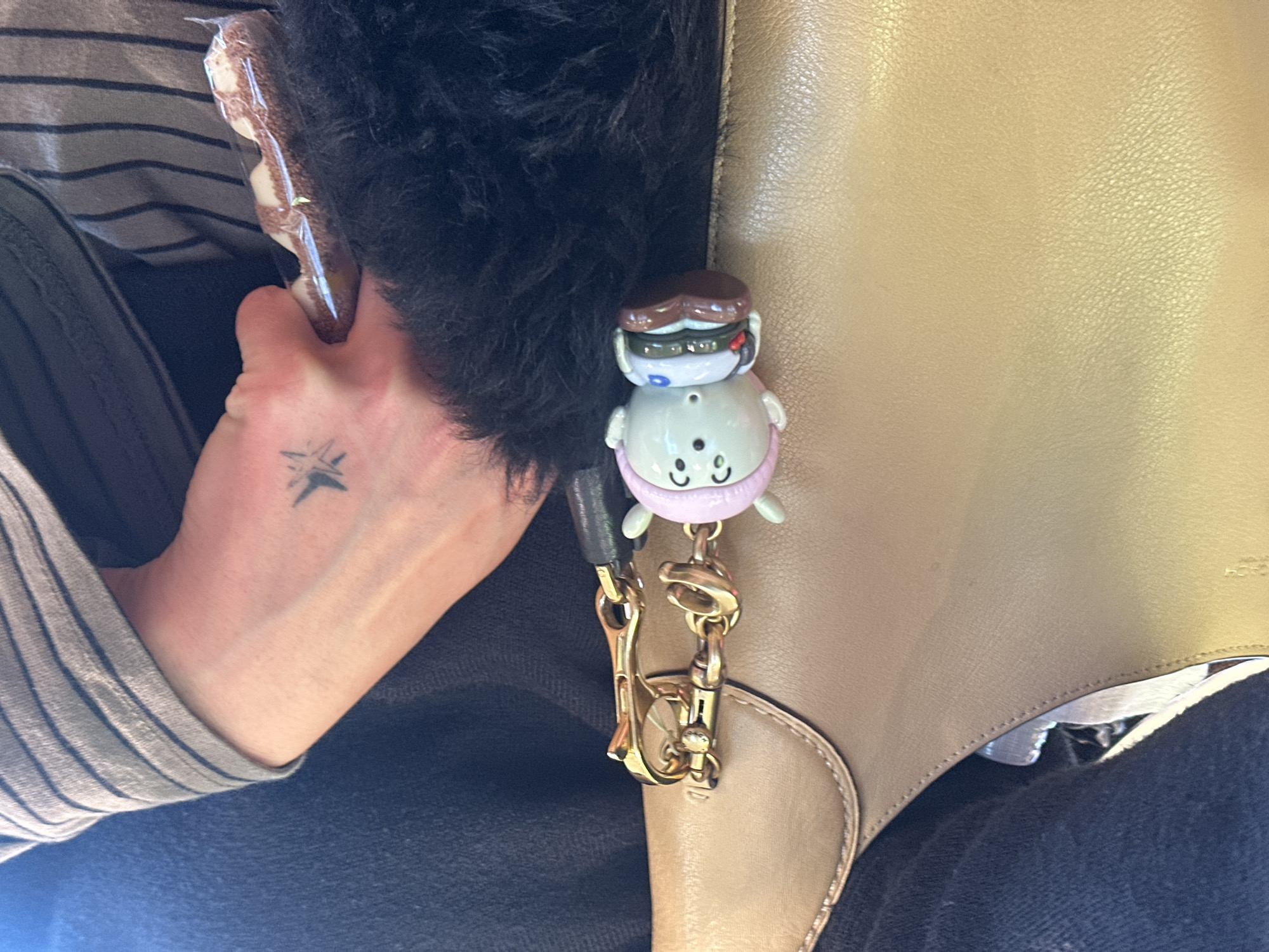 A person's hand, adorned with a small tattoo on the wrist, holds a wrapped snack. The individual is seated, wearing a striped shirt and dark denim jeans. A tan leather bag with a decorative charm is positioned near the person's hand. The scene appears to be indoors, likely within a public setting based on the casual clothing and bag. The exact location in London is not discernible from the image alone.