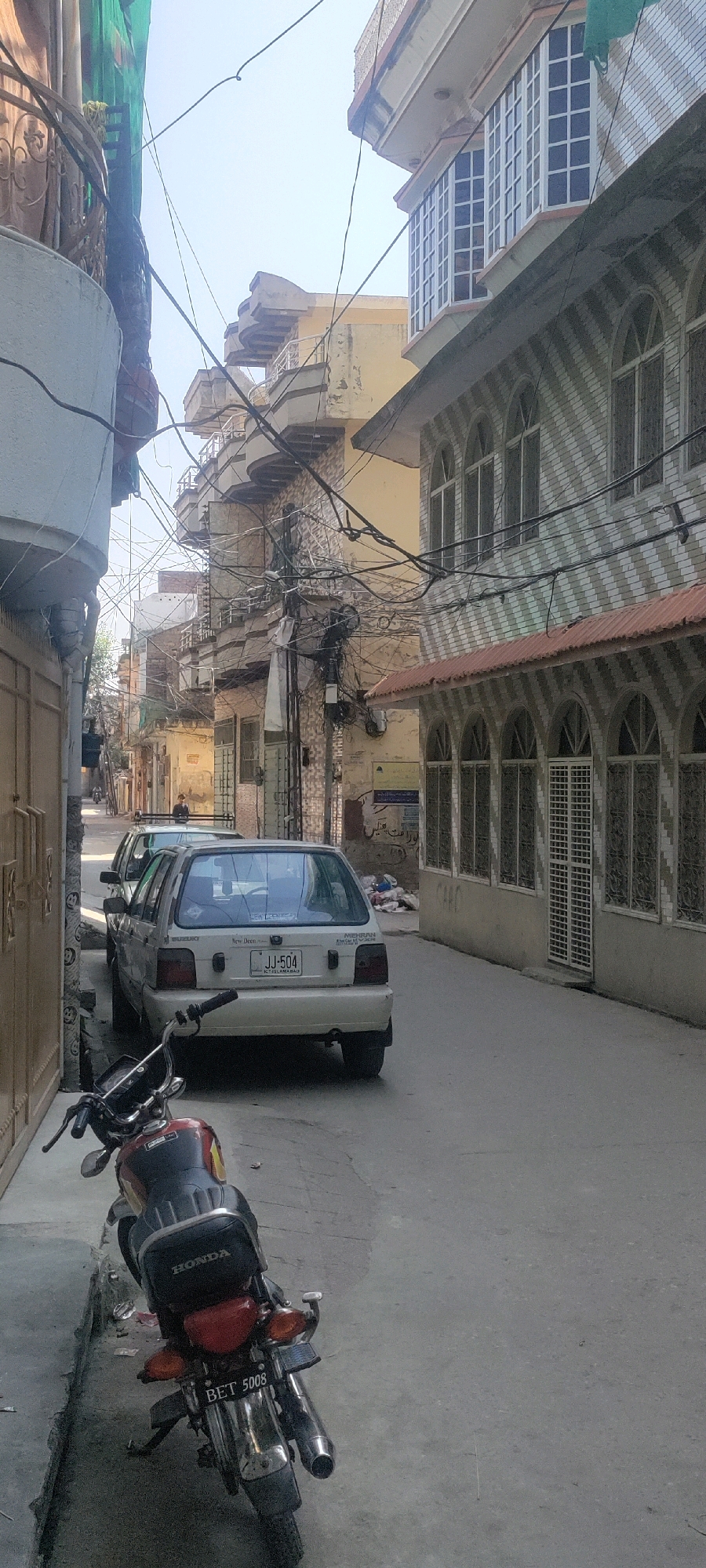 The image depicts a narrow city street in Rawalpindi, Pakistan. In the foreground, a red and black motorcycle with license plate "BET 5008" is parked on the left side of the road. A light-colored Suzuki Mehran car with license plate "JJ-504" is parked further down the street, with another car partially visible behind it. The street is lined with multi-story buildings featuring various architectural styles, including arched windows and ornate facades. Overhead, a dense network of electrical wires crisscrosses the sky, a common sight in Pakistani cities. The scene suggests a typical urban environment with a mix of residential and commercial structures. The lighting indicates it is daytime.