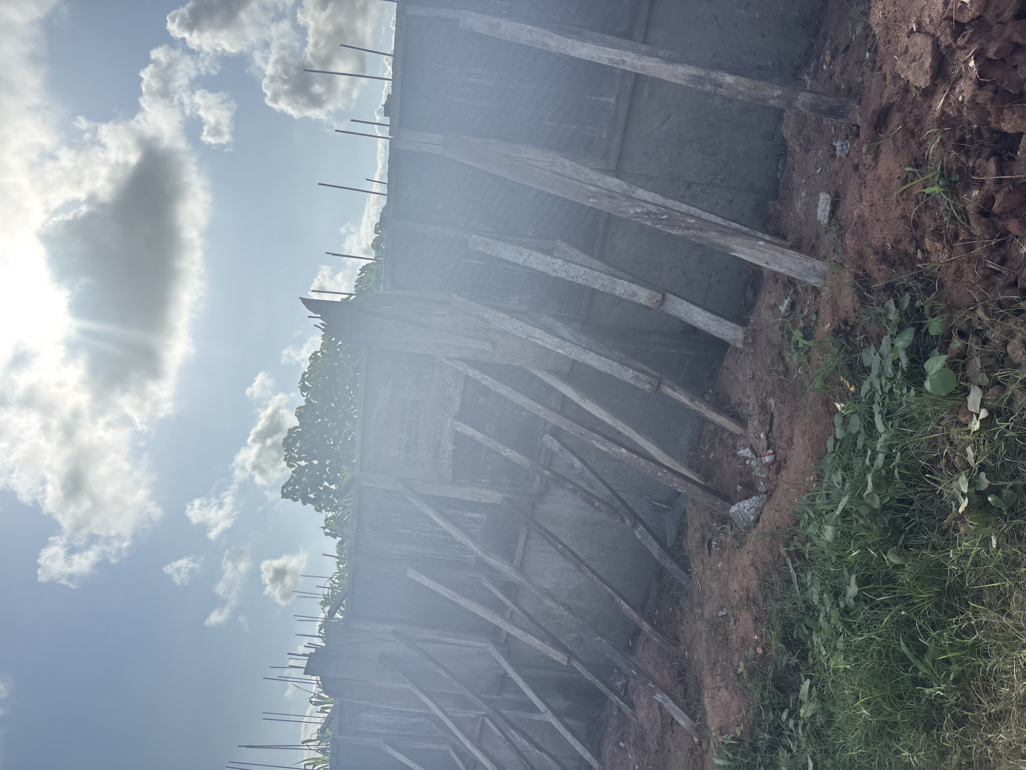 A concrete wall, reinforced with wooden planks, stands tall under a partly cloudy sky. The wall appears to be a construction site, with rebar sticking out from the top. The ground is covered in red soil and patches of green grass. The scene is set in Orifite, Nigeria, under the bright African sun.