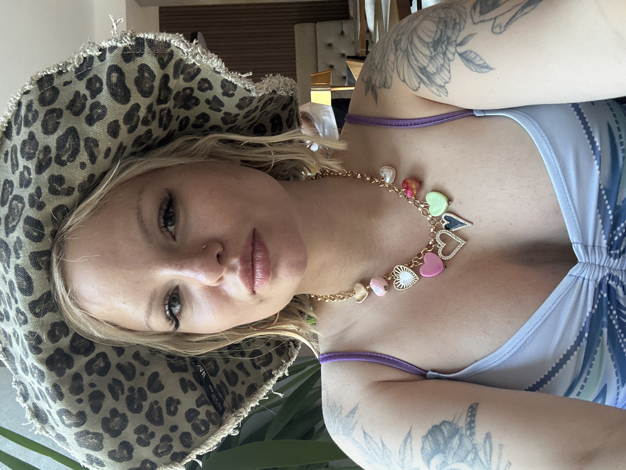 The scene is an indoor setting within the city of Budva, Montenegro. A young woman with shoulder-length blonde hair, light complexion, and a nostril piercing is depicted from the chest up, looking directly at the viewer. She is wearing a leopard print bucket hat with frayed edges, revealing an inverted "ZARA" tag on its brim. Her makeup includes winged eyeliner and mascara, and her lips have a light pink tint. She wears a gold-colored chain necklace adorned with multiple colorful, heart-shaped charms, some solid, some textured, and some encrusted with clear stones. She is dressed in a light-colored top with a subtle pattern and visible purple spaghetti straps. Tattoos are visible on both upper arms, including a large floral/feather design on her right arm. The background on the left features large green plant leaves. On the right, the blurred background shows a warm-toned interior with wooden wall paneling, light-colored upholstered furniture (resembling a tufted headboard), a golden lamp with a rectangular shade, and indistinct figures further in the distance.