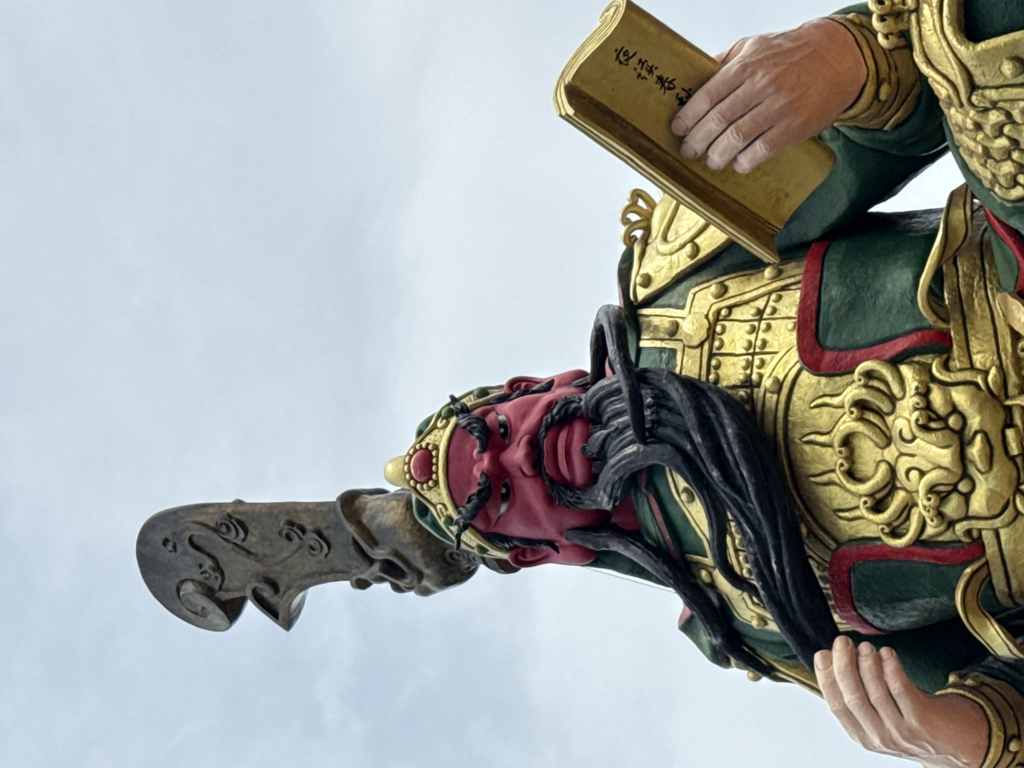 The image shows a low-angle, close-up view of a statue of a warrior, likely a deity or historical figure, located in Baan Hua Thanon, Thailand. The statue depicts a figure with a bright red face, a long black beard, and fierce eyebrows, wearing ornate green and gold armor. A decorative headdress adorns its head. The statue is holding an open, golden book in its right hand, with Chinese characters visible on its pages. The background is a pale, cloudy sky. The statue appears to be made of painted stone or concrete, with intricate detailing on its armor and headdress. The overall impression is one of power and ancient wisdom.
